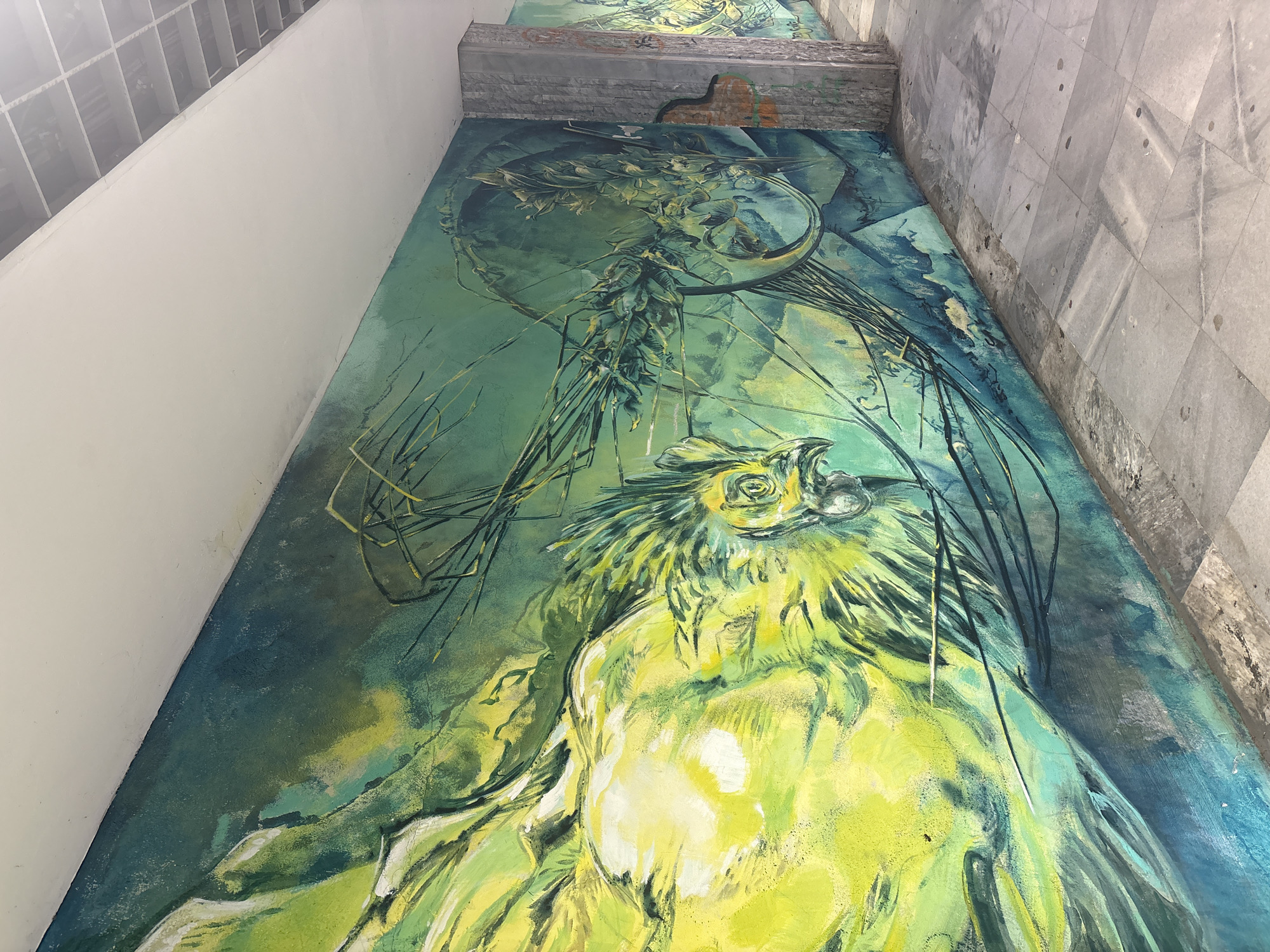 The image shows a mural depicting a bird-like creature in shades of green and yellow. The artwork appears to be painted on a wall surface, possibly in an underground or tunnel-like setting. The mural is positioned adjacent to both a white wall and a grey tiled wall. Above the mural is what appears to be a concrete or stone structure. The mural is located in the city of Unknown, Unknown.