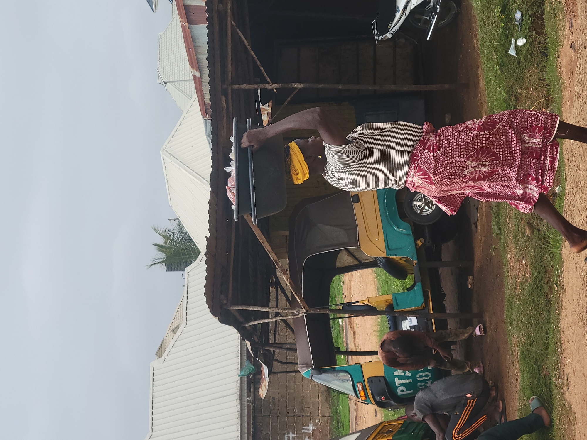 In Barakin Akawo, Nigeria, a woman with a yellow headscarf walks from right to left, carrying two dark rectangular containers stacked on her head, filled with goods. She wears a horizontally striped t-shirt and a long, patterned red skirt. Behind her, a yellow and green auto-rickshaw is parked. A man in a brown shirt and patterned pants is bent over near the auto-rickshaw. To the left, another auto-rickshaw, primarily yellow and green, is partially visible. In the background, a row of buildings with corrugated metal roofs stands under an overcast sky. A weathered, open-air stall with a corrugated metal roof is situated in front of some of the buildings, supported by rough wooden poles. A white and black motorcycle is parked to the right of the stall. The ground is a mix of dirt and short green grass.