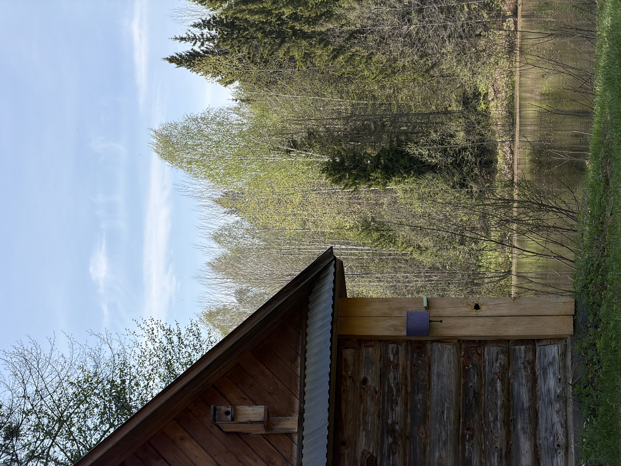 The image shows a rustic wooden building with a steeply pitched roof covered in corrugated metal. A small birdhouse is attached to the side of the building near the roofline. Adjacent to the building, there is a view of a lake or pond surrounded by trees, including evergreens and deciduous trees with budding leaves. The sky is partially visible and appears to be lightly cloudy. The scene appears to be rural and wooded. The specific location is Ural'skoe, Russia. There is no indication of people or interaction shown in the photo.
