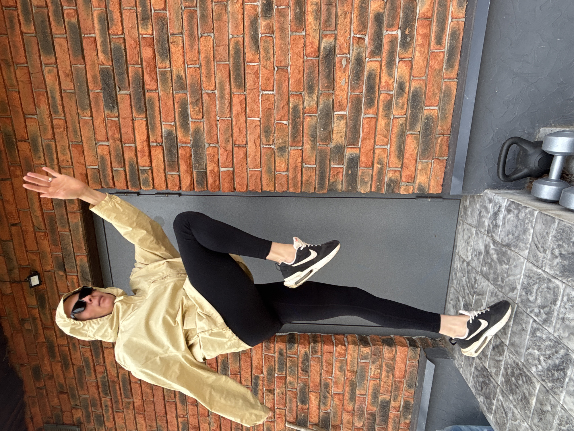 The image features a person, likely female, in a dynamic, athletic pose against a textured background. The individual is wearing a light khaki or beige hooded windbreaker jacket, black leggings, and black Nike athletic shoes with white soles and white swoosh logos. Dark sunglasses obscure their eyes. A silver ring is visible on the ring finger of their left hand, which is extended.
The person's pose suggests movement or a balanced stance, with their left arm raised, right arm slightly bent, and one leg bent at the knee while the other is extended downwards, planted on a grey surface.
The background consists of a prominent brick wall, characterized by a pattern of reddish-orange bricks interspersed with darker, almost black, bricks, arranged in vertical columns. Below the brick wall, a dark grey floor or platform is visible. To the right of the person, on a lighter grey, possibly tiled, surface, gym equipment is present, including a black kettlebell and two silver dumbbells.
The bright, natural lighting indicates that the photo was taken during the daytime. The setting combines architectural elements with fitness equipment, suggesting an outdoor or semi-outdoor exercise area in Chaikovsky, Russia. The only visible text is the Nike brand logo on the shoes.