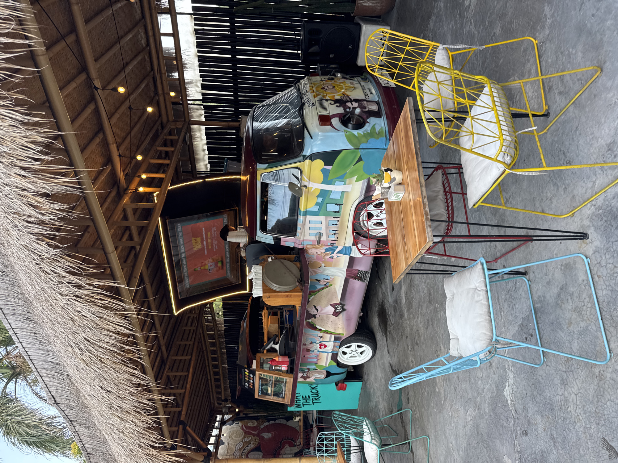 The image displays a vibrant and casual outdoor dining or food service area, characteristic of a tropical setting in Kuta, Indonesia. The central feature is a vintage-style food truck, intricately painted with colorful, cartoon-like murals depicting various characters, landscapes, and food items, such as a large sandwich. The truck's body is a lively mix of blue, yellow, purple, and red.
Surrounding the food truck are eclectic metal wire chairs in bright yellow and light blue, each furnished with a white cushion, arranged around a rustic wooden table. The floor is a weathered concrete. Overhead, a thatched roof, likely made of palm fronds and supported by bamboo beams, provides shelter. Warm-toned string lights are strung across the ceiling, suggesting an evening ambiance or a shaded environment. Palm fronds are visible on the left side, reinforcing the tropical location. Black speakers are positioned near the top right, hinting at a lively atmosphere.
The primary text visible includes "WHAT THE TRUCK," prominently displayed on the vehicle itself and on a nearby sign. Other text, possibly from a menu or promotion, reads "DO WHAT THE TRUCK WHAT THE TRUCKT WHAT THE TRUCK," "HAPPY HOU," and an implied "BUY 1 GET 1 FREE" (from "BUY I GET HIL"). The scene implies a relaxed gathering place for food and drinks, though no people are distinctly visible.
