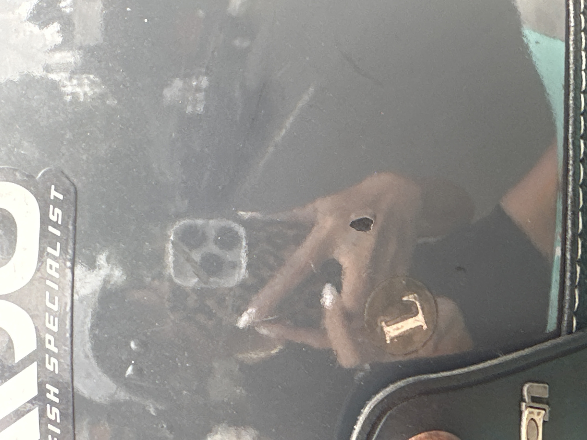 The image depicts a reflective dark grey surface, possibly a vehicle panel or equipment, located in Batubulan, Indonesia. The surface exhibits a small, irregular dark blemish or hole in the middle-right area. Text elements are visible on the surface: partially cropped white letters "KSO" are in the upper left, and "FISH SPECIALIST" in white text is positioned below them. In the lower left, a dark, rounded component features partially visible metallic letters "AN". A circular sticker bearing the white letter "L" is affixed to this component.
Reflected in the surface is a person taking the photograph. The person has dark, possibly long hair, and is wearing dark sunglasses with roundish frames. A dark top or dress is visible on their torso, and a light-colored ring is discernible on their left ring finger. Their fingernails are painted white or a light color. The person is holding a dark-cased smartphone, which is clearly reflected, revealing a distinct square camera module with multiple lenses and what appears to be a flash or sensor array. Additionally, reflections indicate a bright, light-colored area in the upper right, and a teal or light blue object in the lower right, suggesting an outdoor environment. The reflective surface itself appears speckled with dust or debris.
