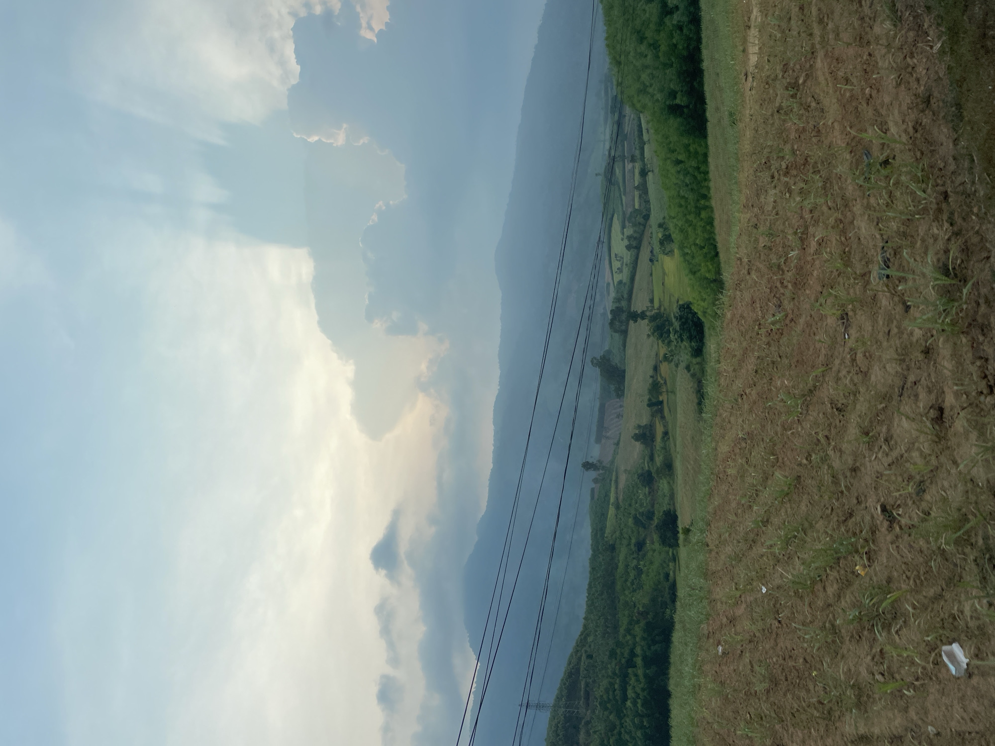 The image shows a landscape in Thôn 18, Vietnam. The sky is partially cloudy, with light blue patches and large, billowing clouds. A mountain range fades into the distance, obscured by haze or mist. Power lines stretch across the scene, leading towards the mountains. Patches of green fields and trees are interspersed among areas of cultivated land. In the foreground, there is a field with dry grass and sparse vegetation. Some scattered debris, possibly plastic or paper, is visible on the ground. The image is taken at an angle, with the camera likely tilted to the side.