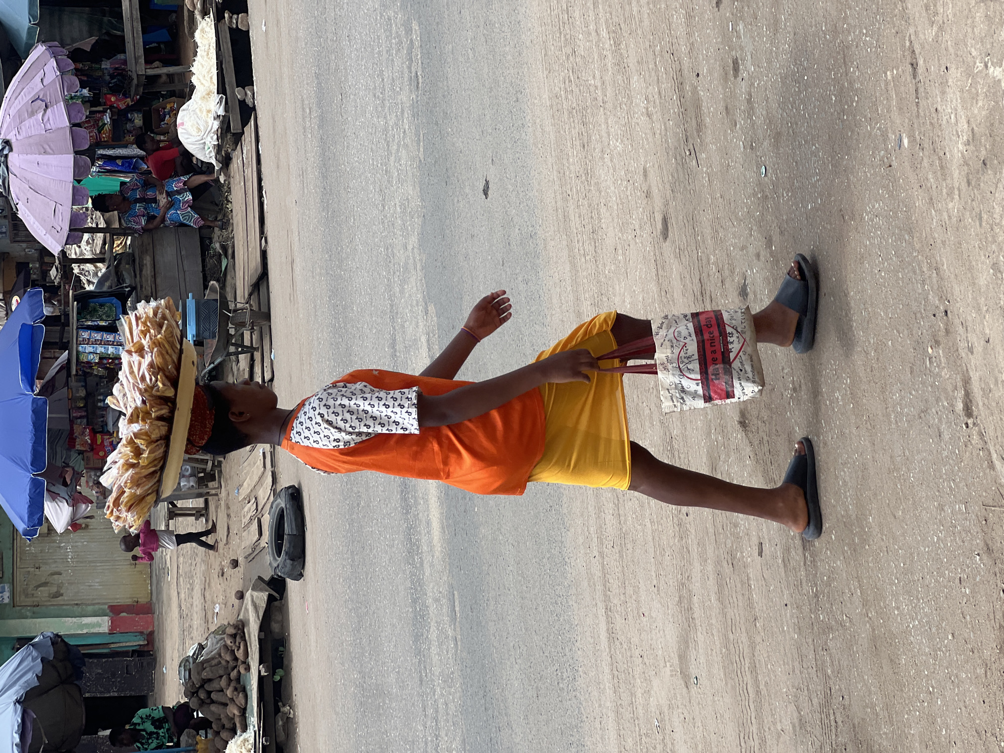 A young individual is walking on a paved street in Nkpor, Nigeria, carrying a large tray of food on their head. The tray appears to contain packaged snacks. The individual is wearing an orange shirt, yellow shorts, and black sandals. They also carry a shopping bag in their hand. The background includes market stalls with various goods, including yams and other food items. There are umbrellas shading the stalls. Other people are present in the background, including vendors and shoppers. A tire lies on the ground near the stalls.