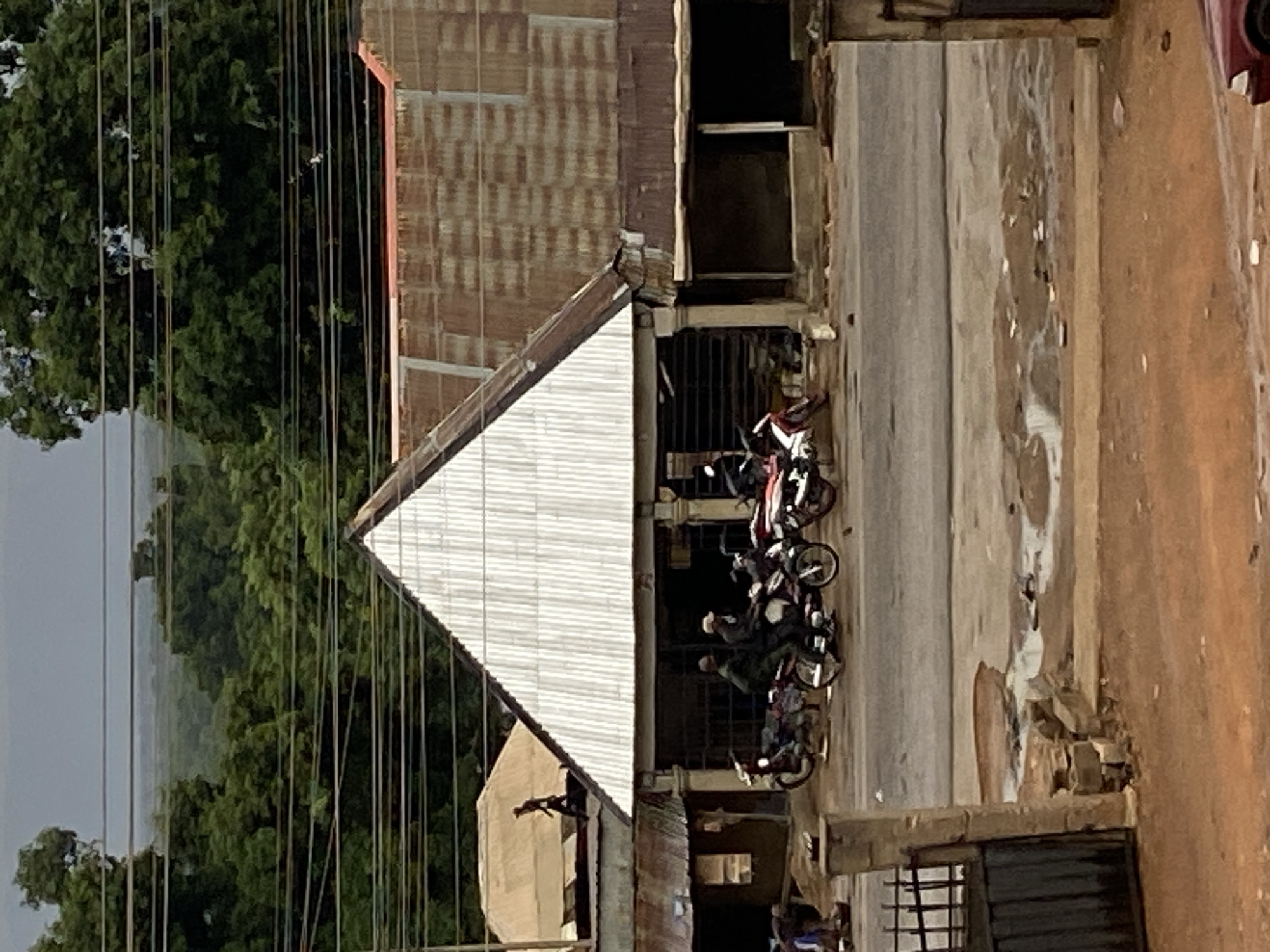The image depicts a street scene in Mararaba, Nigeria. Several motorcycles are parked along the side of the road near buildings with corrugated metal roofs. Lush green trees are visible in the background, along with utility lines. The street surface appears to be concrete or paved. The overall lighting suggests it is daytime.