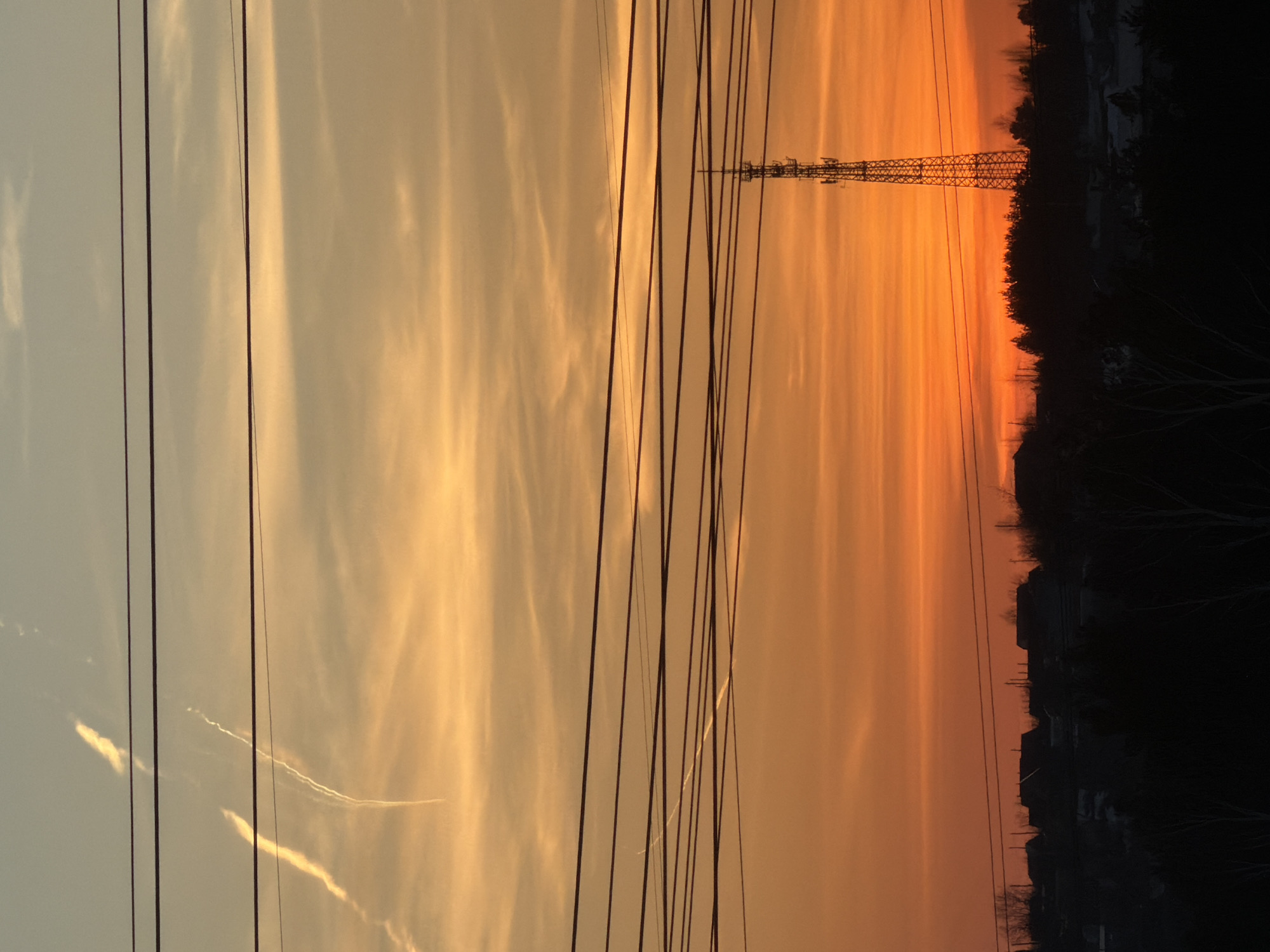 The image depicts a sunset in what appears to be the city of Chaikovsky, Russia, as evidenced by the power lines and the silhouette of a lattice tower possibly associated with communication or power transmission infrastructure. The sky exhibits a gradient of color from a lighter, yellowish hue to a deeper orange, suggesting the sun is setting or rising. Silhouetted against the brighter sky is a treeline and what seems to be a residential or industrial area. There is no evidence of people or direct interaction in the scene. The general environmental context is an urban or suburban landscape at twilight. There appear to be contrails from an airplane in the sky. The image is vertically oriented.