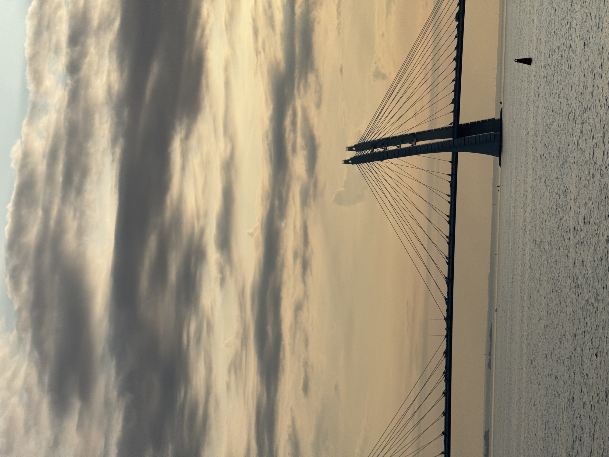 A long-exposure shot of the Gulf of Finland at sunset, with the Western High-Speed Diameter bridge silhouetted against a cloudy sky. The water is calm, with gentle waves, and a small red buoy floats in the foreground. The bridge's distinctive A-frame pylons are visible, along with the suspension cables stretching across the frame. The sky is filled with layered clouds, ranging from light gray to a warm, golden hue, indicating the setting sun. In the distance, the faint outline of the St. Petersburg skyline is visible.