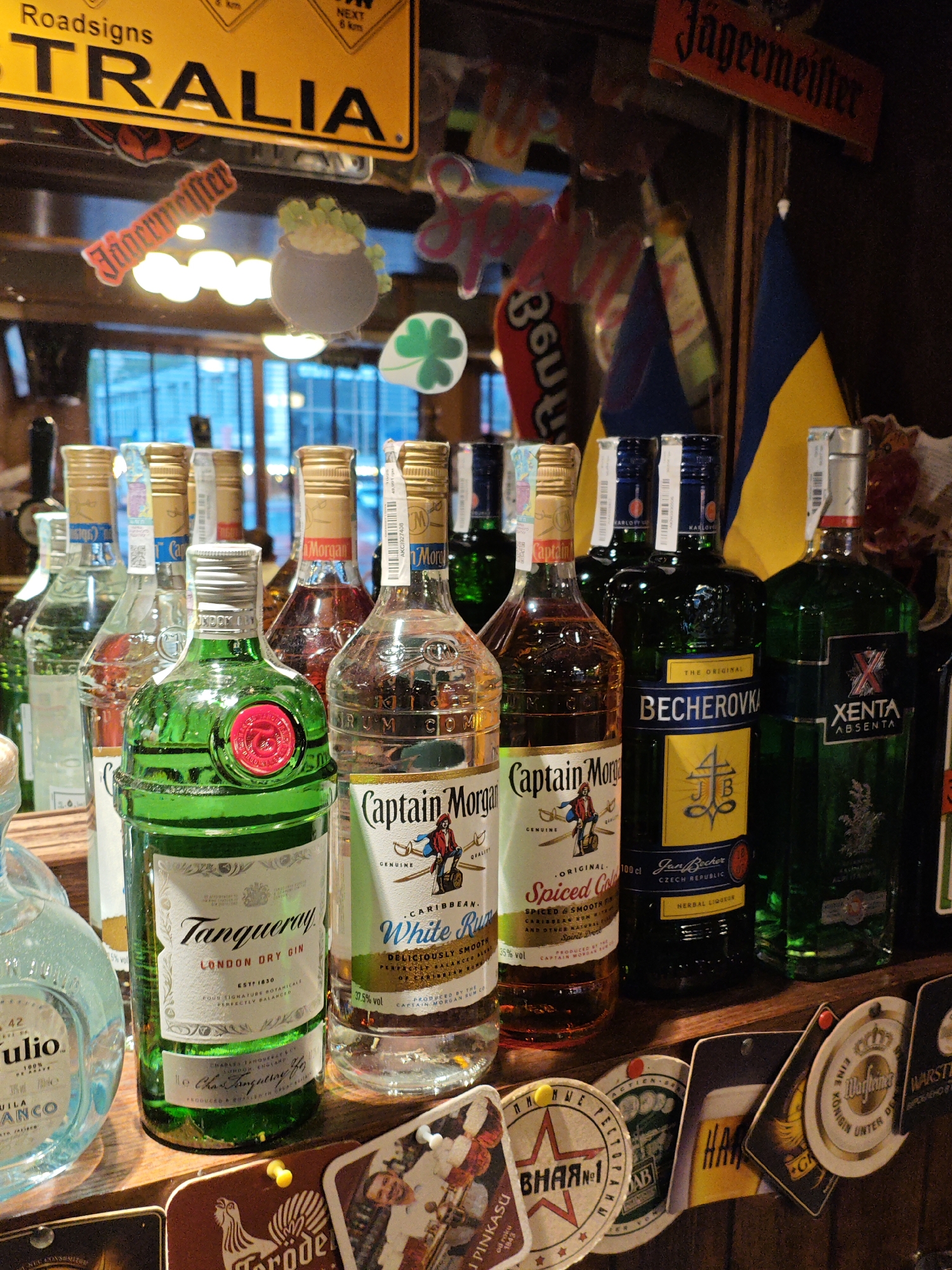 The image shows a close-up, eye-level view of a bar shelf filled with various bottles of alcohol. In the foreground, prominently displayed, is a green bottle of Tanqueray London Dry Gin, a bottle of Captain Morgan Spiced Gold Rum, and a bottle of Captain Morgan White Rum. Behind these, several other bottles are visible, including a dark green bottle labeled "Becherovka" and a green bottle with a red "X" logo, identified as "Xenta Absenta." The background features a collection of flags, including a Ukrainian flag, and various decorative elements like a sign that says "Roadsigns TRALIA" and a smaller sign that says "Jagermeister." The lighting is dim, characteristic of a bar environment, with some reflections visible on the bottles and the shelf. Several beer coasters are also scattered on the shelf in front of the bottles. No people are visible in the image. The scene is set in a bar, likely in a city like Kyiv, Ukraine, suggested by the presence of the Ukrainian flag and the overall ambiance.
