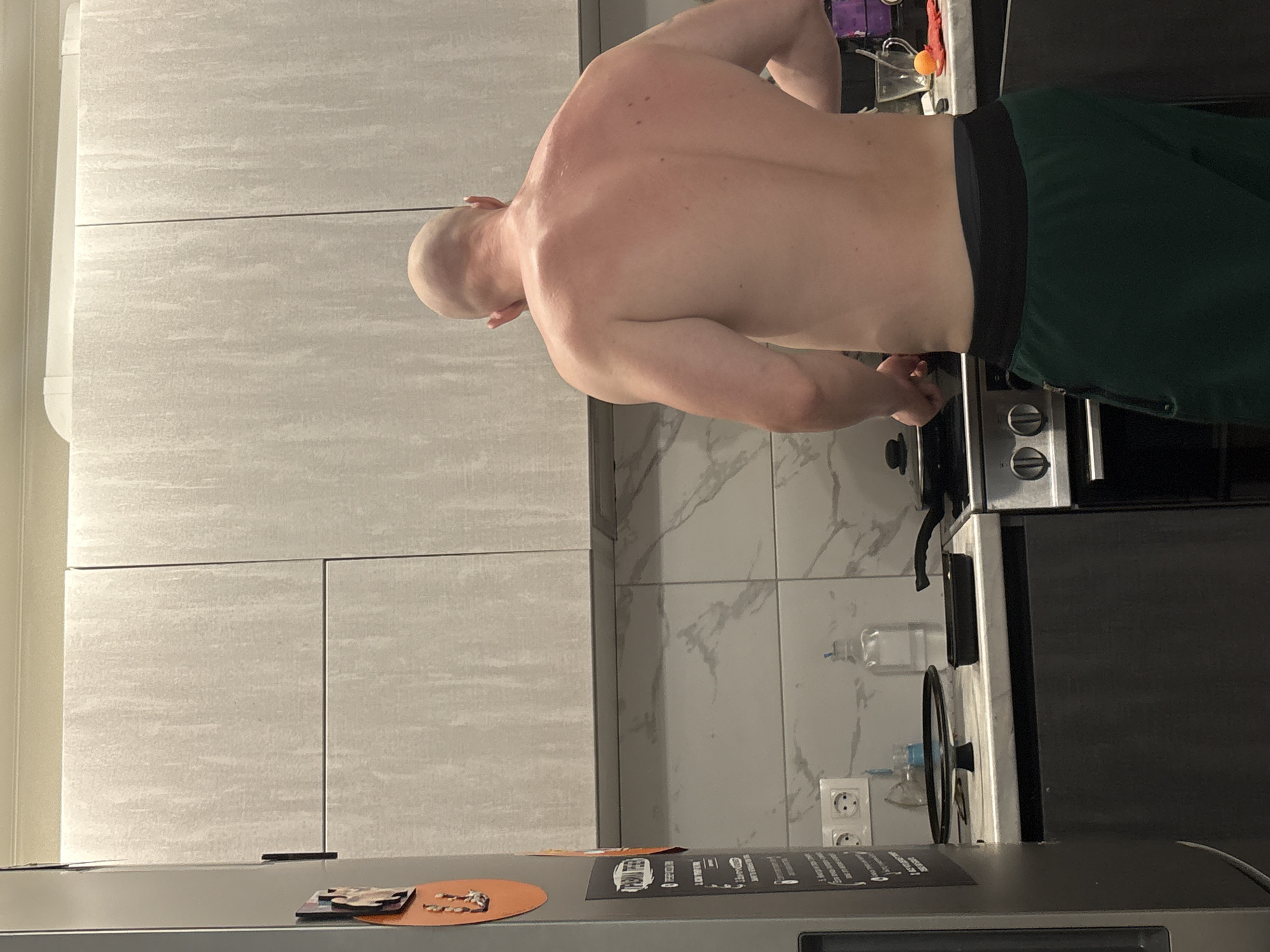 A shirtless man with a shaved head stands in a kitchen. He wears dark green shorts with a black waistband. His back is to the camera, and he appears to be interacting with something on the stove. The kitchen features light grey cabinets and a stove with silver-colored knobs. There is a marble-patterned tile backsplash behind the stove. A refrigerator is visible in the foreground. The image does not give enough information to verify the location as Izhevsk, Russia.