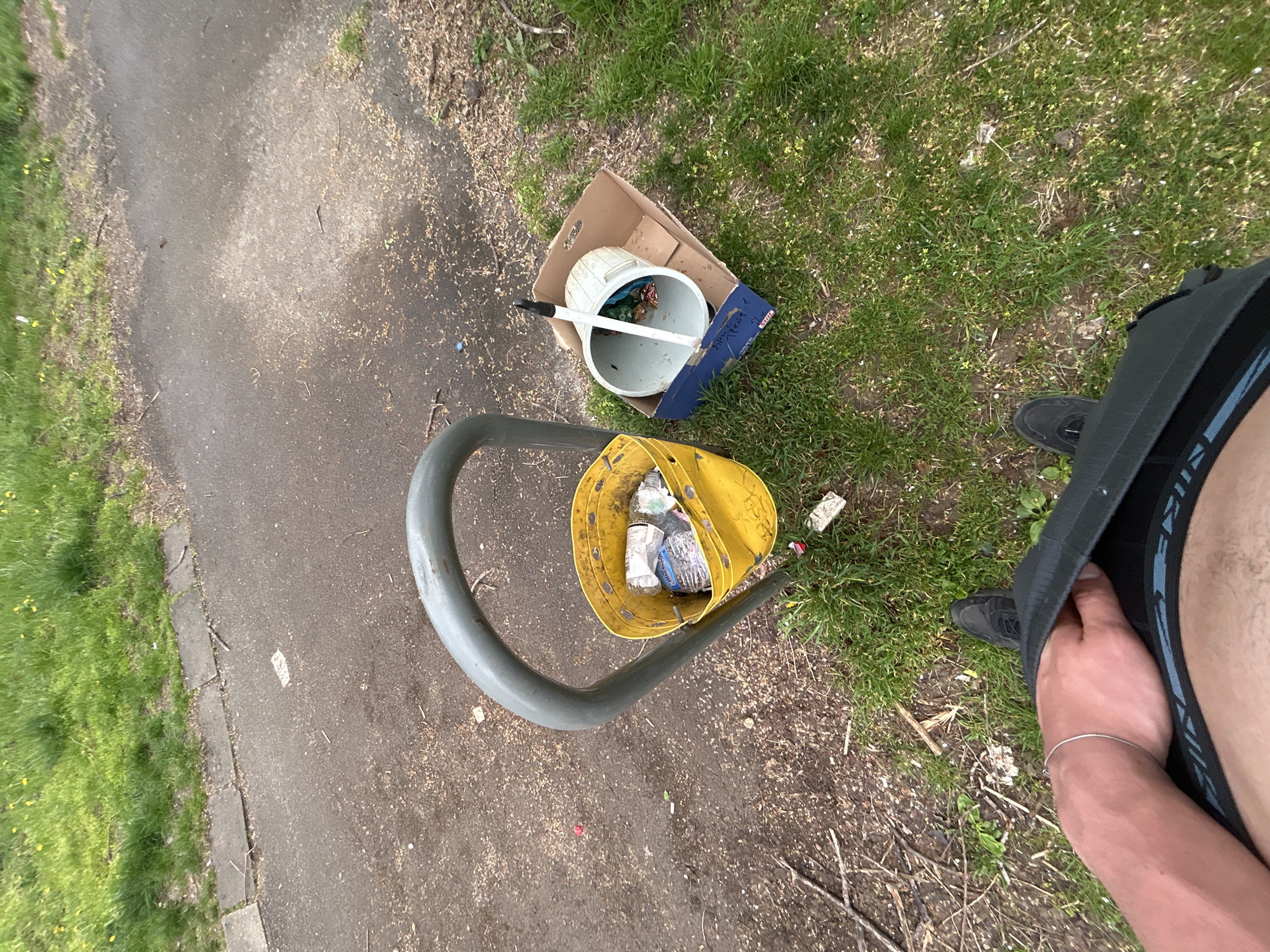 A top-down shot taken outdoors depicts two buckets containing refuse, positioned on grass and a paved walkway, presumably in a public space. A yellow bucket filled with plastic bottles and other trash hangs from a gray metal bike rack. A cardboard box nearby holds a white bucket containing a mop or broom. The right edge of the image shows a person's right leg and torso, their hand covering their lap, and one foot visible with a black shoe. The setting appears to be near a grassy area with a paved path, suggestive of a park or recreational area. Without further context, it is impossible to confirm this location to be in Sofia, Bulgaria.