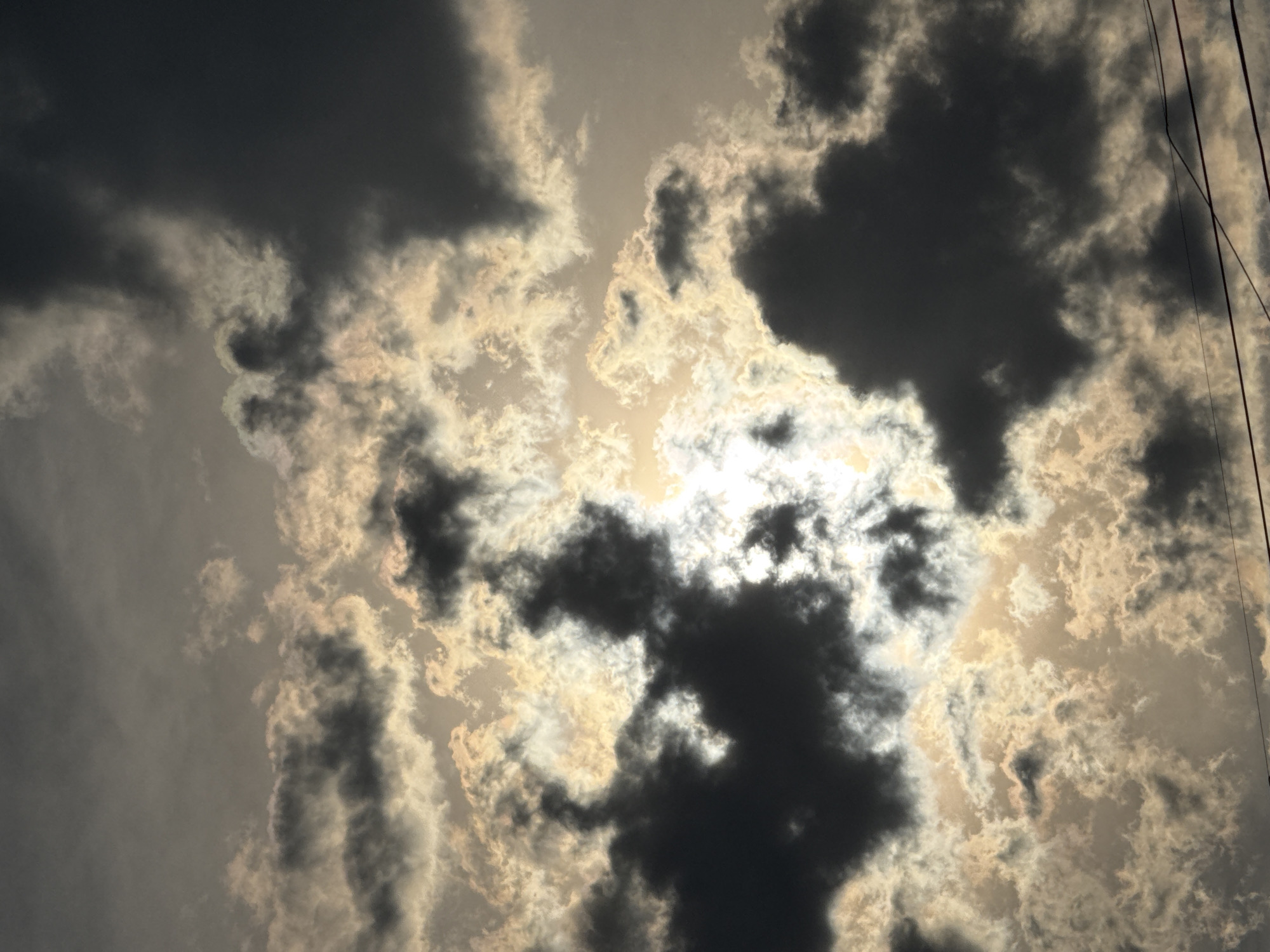 The image depicts a dramatic sky filled with dark, billowing clouds illuminated by a bright, partially obscured sun. The sunlight filters through the clouds, creating a contrast between light and shadow and casting an almost ethereal glow on some of the cloud formations. In the lower portion of the image, a series of utility wires are visible, stretching horizontally across the frame, suggesting an urban or semi-urban setting. The overall mood is somber yet awe-inspiring, characteristic of a turbulent weather system. There are no people or specific structures visible in this image. The location is identified as Abovyan, Armenia.