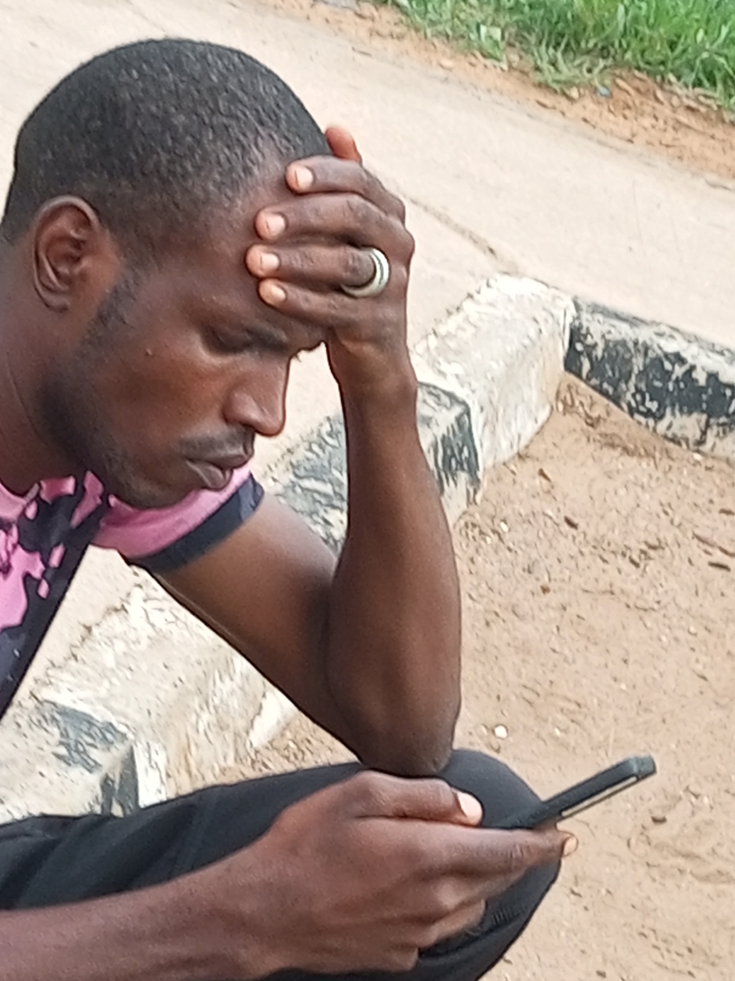 A man with dark skin and short black hair is seated outdoors, his right hand resting on his forehead and left hand holding a black smartphone. He is wearing a pink shirt with black accents and dark pants. The man appears to be looking down at the phone. He has a ring on the ring finger of his right hand. The background consists of a dirt path, a concrete curb, and some green vegetation. The scene is set in the city of Gombe, Nigeria.