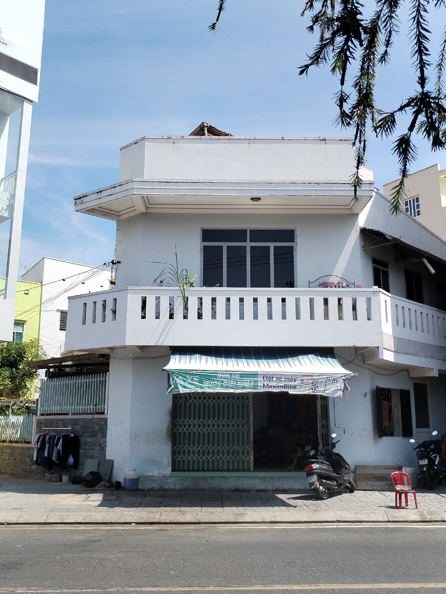 The image captures a street view of a white, multi-story building in Nha Trang Ward, Vietnam. The building appears to be a commercial establishment on the ground floor, with a green metal gate covering the entrance. A striped awning is stretched over the entrance, displaying text that reads "Dat xe may MaximBike". Two motorcycles are parked in front of the building, and a red plastic chair sits on the sidewalk. Clothes are hanging on a rack to the left of the entrance. The building has a white balcony with decorative railings on the second floor, overlooking the street. The sky is clear and blue, with tree branches hanging into the frame from the top right corner. The scene is illuminated by bright sunlight, casting shadows on the ground.