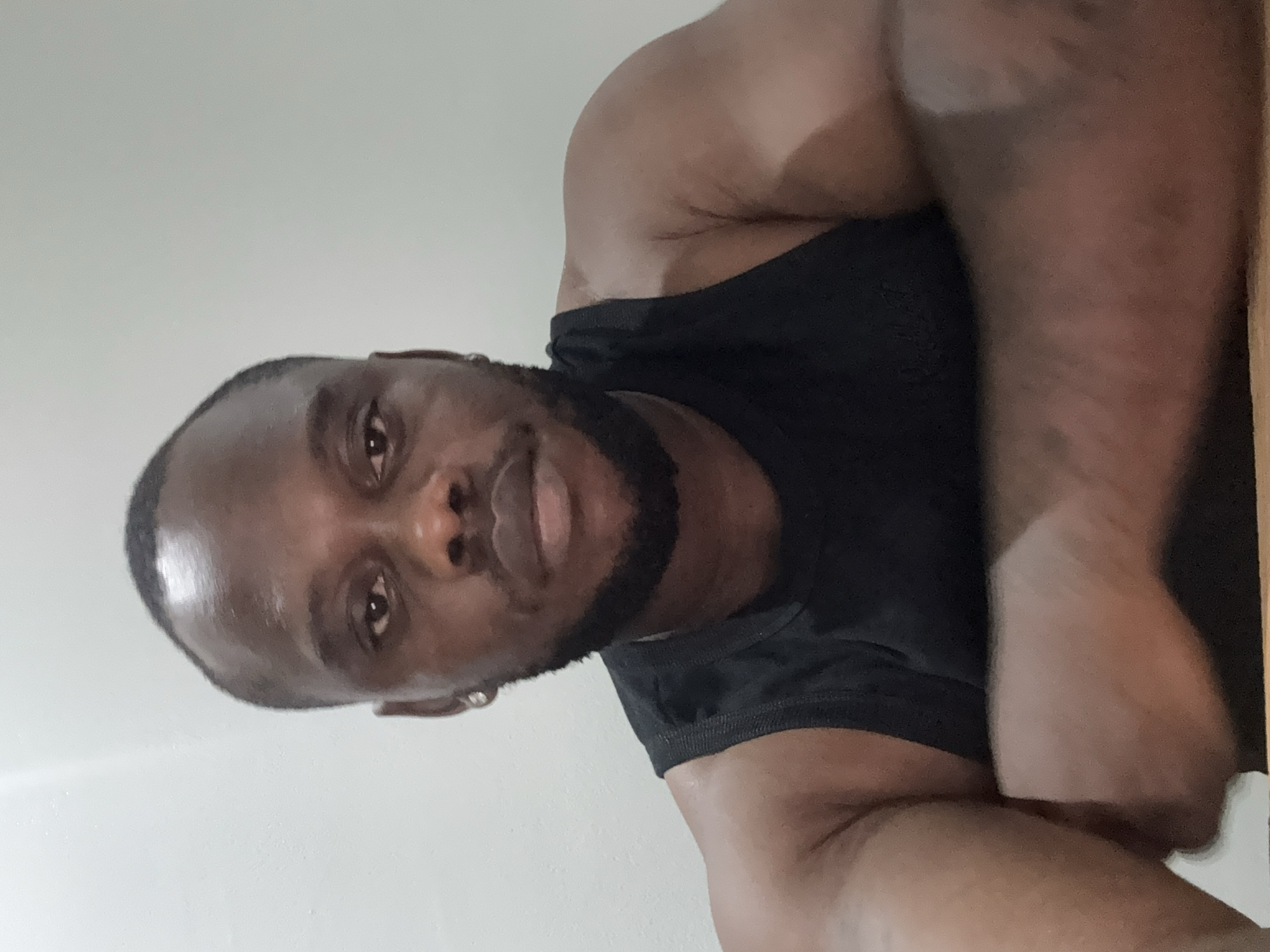 A portrait-style image, rotated 90 degrees clockwise, features an adult man with dark skin. He has short, dark hair and a neatly trimmed short beard or stubble. He is wearing a black sleeveless top or tank top, revealing muscular arms. A small, silver stud earring is visible in his left ear. The man looks directly at the camera with a neutral to slightly soft expression, and his gaze is direct and focused.
The background consists of a plain, light-colored wall on the left side of the frame, and a darker, possibly wooden or textured surface on the right side. The man appears to be in an indoor setting. The lighting is even, suggesting an indoor light source, and there are no specific visual cues to indicate the time of day or weather. No text is visible in the image.