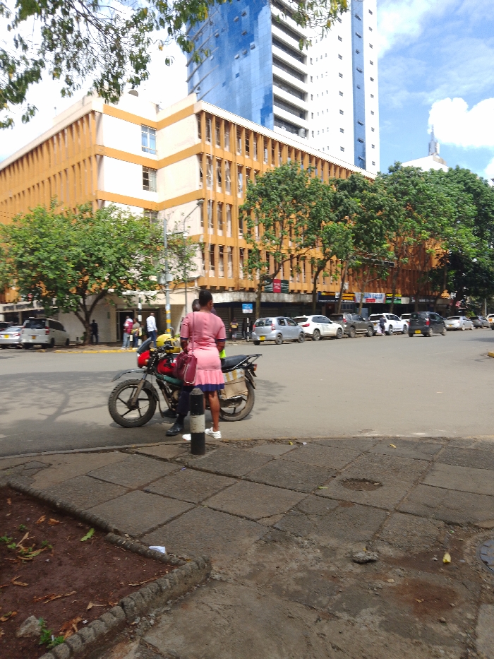 The image shows a street corner in Athi River, Kenya, at the intersection of a paved road and a sidewalk. In the foreground, there is a parked motorcycle with a woman standing next to it. She is wearing a pink dress and carrying a pink purse. She is speaking to a man who is wearing a yellow helmet and a black jacket. The man is holding a yellow helmet. The sidewalk is made of large concrete slabs. In the background, there is a street lined with parked vehicles. The street is busy with cars and pedestrians. The street is lined with trees and buildings, including a tall, modern office building. The building is painted blue and has many windows. It is located on the left side of the image. There are several other buildings in the background, including a yellow building with a large sign. The sky is clear and blue with a few clouds. The scene is situated in Athi River, Kenya, at the intersection of a paved road and a sidewalk.