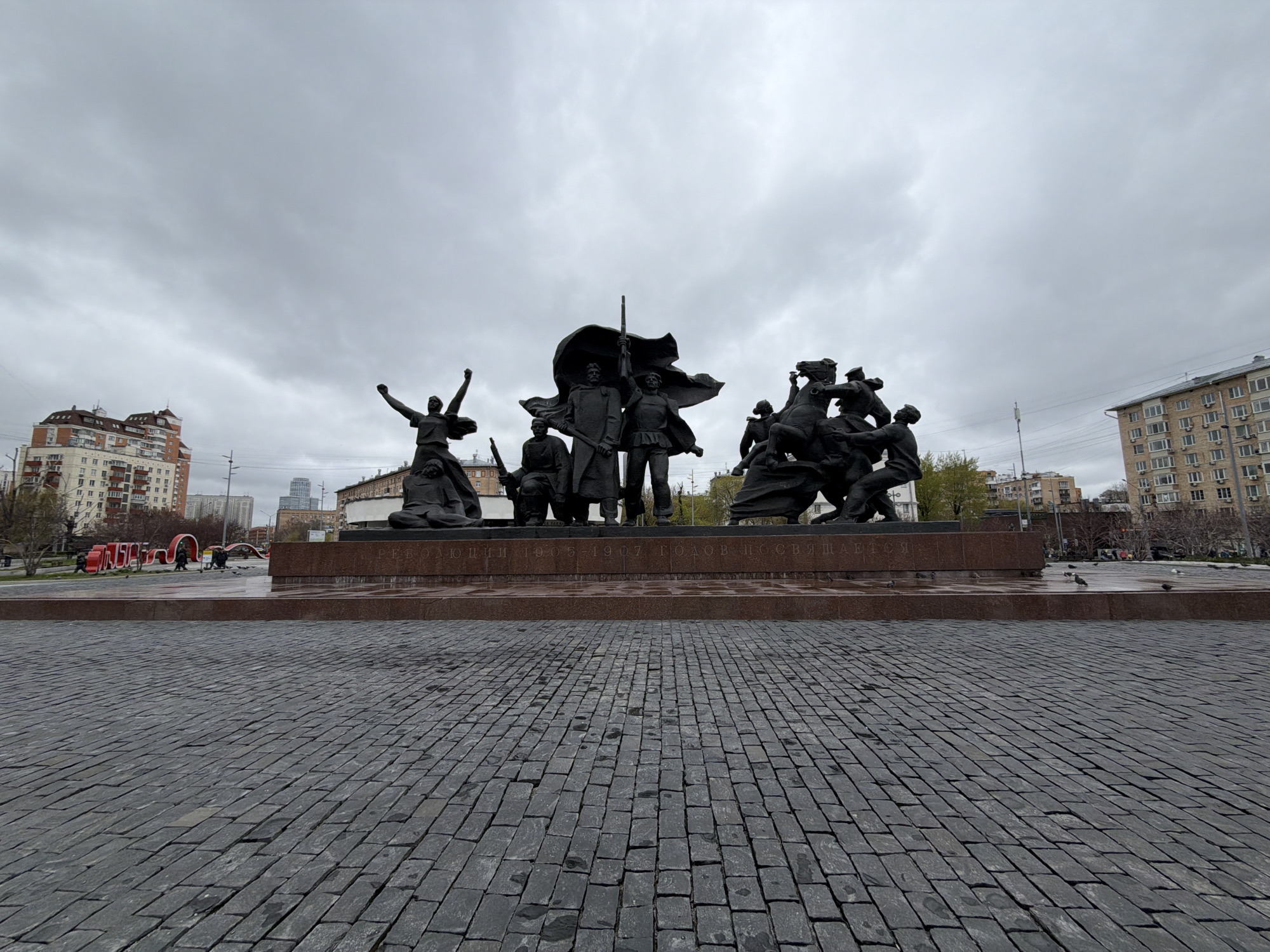 This wide-angle shot features a monument in Moscow, Russia dedicated to the Revolution of 1905-1907. The monument depicts a group of figures, including a woman with raised arms, several men with a flag and rifles, and a group of horsemen. The monument is placed on a raised platform with text in Cyrillic script. The foreground is composed of a gray cobblestone pavement that stretches towards the base of the monument. In the background, there are several buildings, including a brick-colored building with multiple levels and a building with a light beige facade and rows of windows. The sky is overcast.