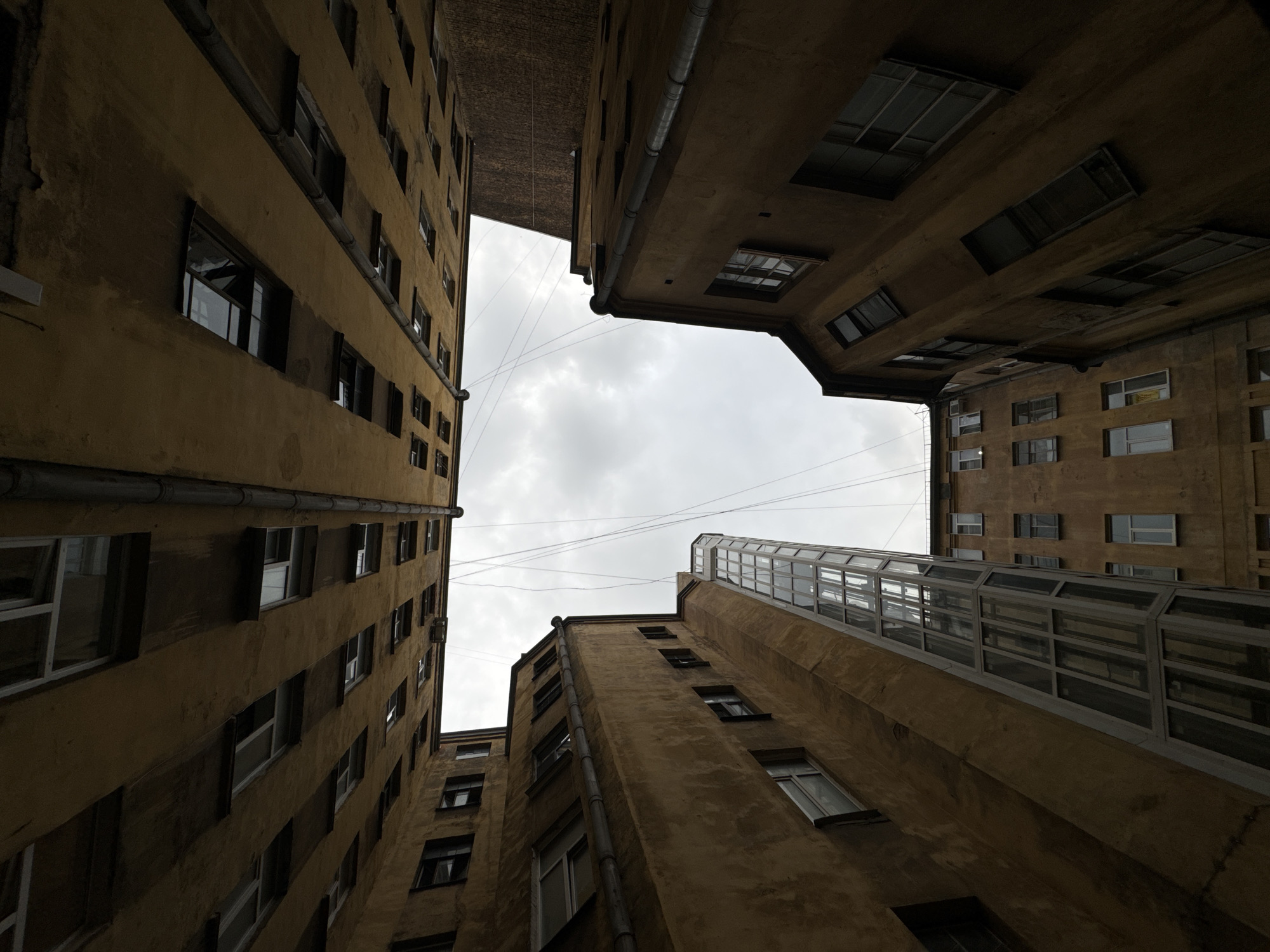 This is a low-angle shot of a narrow courtyard in Saint Petersburg, Russia, looking upwards towards a cloudy sky. The courtyard is enclosed by tall, old buildings with weathered yellow facades. The buildings have numerous windows, some open and some closed, arranged in neat rows. A modern glass elevator enclosure is attached to one of the buildings, contrasting with the older architecture. Thin wires crisscross the sky between the buildings. The overall impression is one of enclosed urban space, with a sense of history and decay.