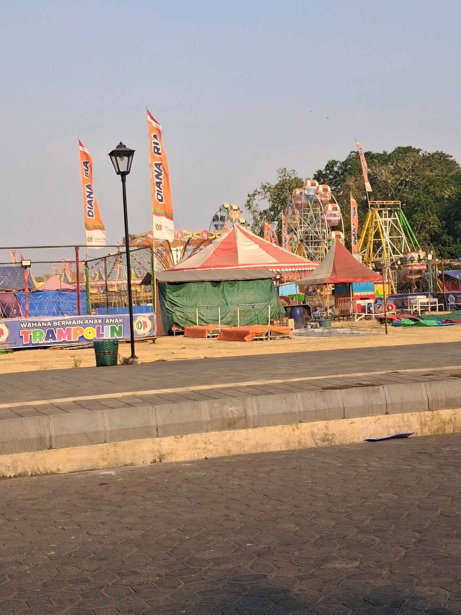 A medium-shot outdoor photograph shows an amusement park in the city of Surakarta, Indonesia. In the foreground, a patterned pavement and concrete border are visible. Beyond the border is a dirt path. In the background, several amusement rides are present, including a Ferris wheel and a swing ride. A trampoline attraction is also visible, with a banner that reads "WAHANA BERMAIN ANAK-ANAK TRAMPOLIN". Two flags with the text "DIANA RIA" are planted in front of a lamppost. The sky is clear and a few birds are flying in the distance. The overall atmosphere is that of a sunny day at an amusement park.