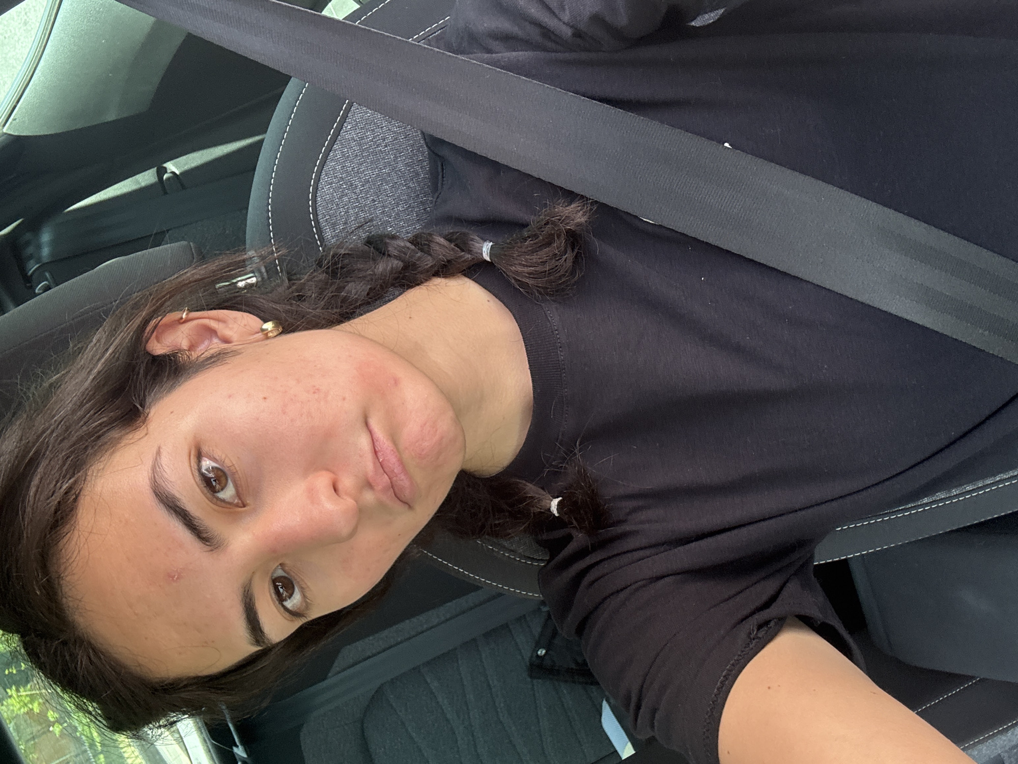 A person wearing a black shirt and a seatbelt is sitting in a car. The person has brown hair, styled in a braid, and brown eyes. The car's interior is visible, featuring grey upholstery with white stitching. The location is not discernable as being in San Bernardino Tlaxcalancingo, Mexico, based on the visual information present in the image.