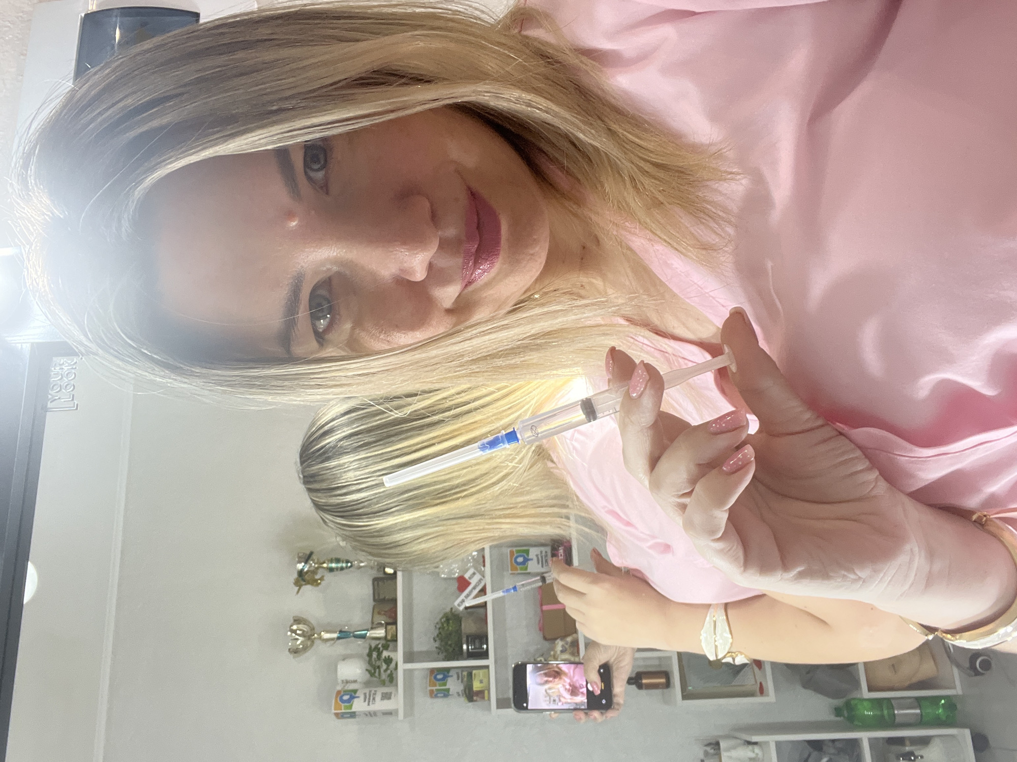 A blond woman with light skin and pink lipstick is captured in a mirror selfie, holding a syringe in her right hand and a smartphone in her left. She wears a pink, possibly satin, top or robe. Her hair is styled in a shoulder-length cut with lighter highlights. Her eyes are light-colored, and she has a slight, calm smile.
In her right hand, she holds a transparent syringe filled with a clear liquid, which is pointed towards the viewer. Her fingernails are manicured with light pink polish. In her left hand, she holds a black smartphone, which is actively displaying a live feed of her reflection, showing that she is taking the photo. Several bracelets, including gold-toned and white shell-like designs, adorn her left wrist.
The setting appears to be an indoor room, possibly a personal space or a professional treatment room, in Sochi, Russia. The background, visible in the mirror reflection, features white shelving units. These shelves hold various items, including small golden trophies or decorative pieces, several bottles and jars, and a green plastic bottle on a lower shelf. Visible text on some of the items includes "100 RASPBERRY" on a box and a distinctive "Q" logo within a circle on multiple product containers. The edge of the mirror frame is also partially visible, showing faint, partially obscured text such as "LOG". The overall lighting suggests an indoor, well-lit environment.