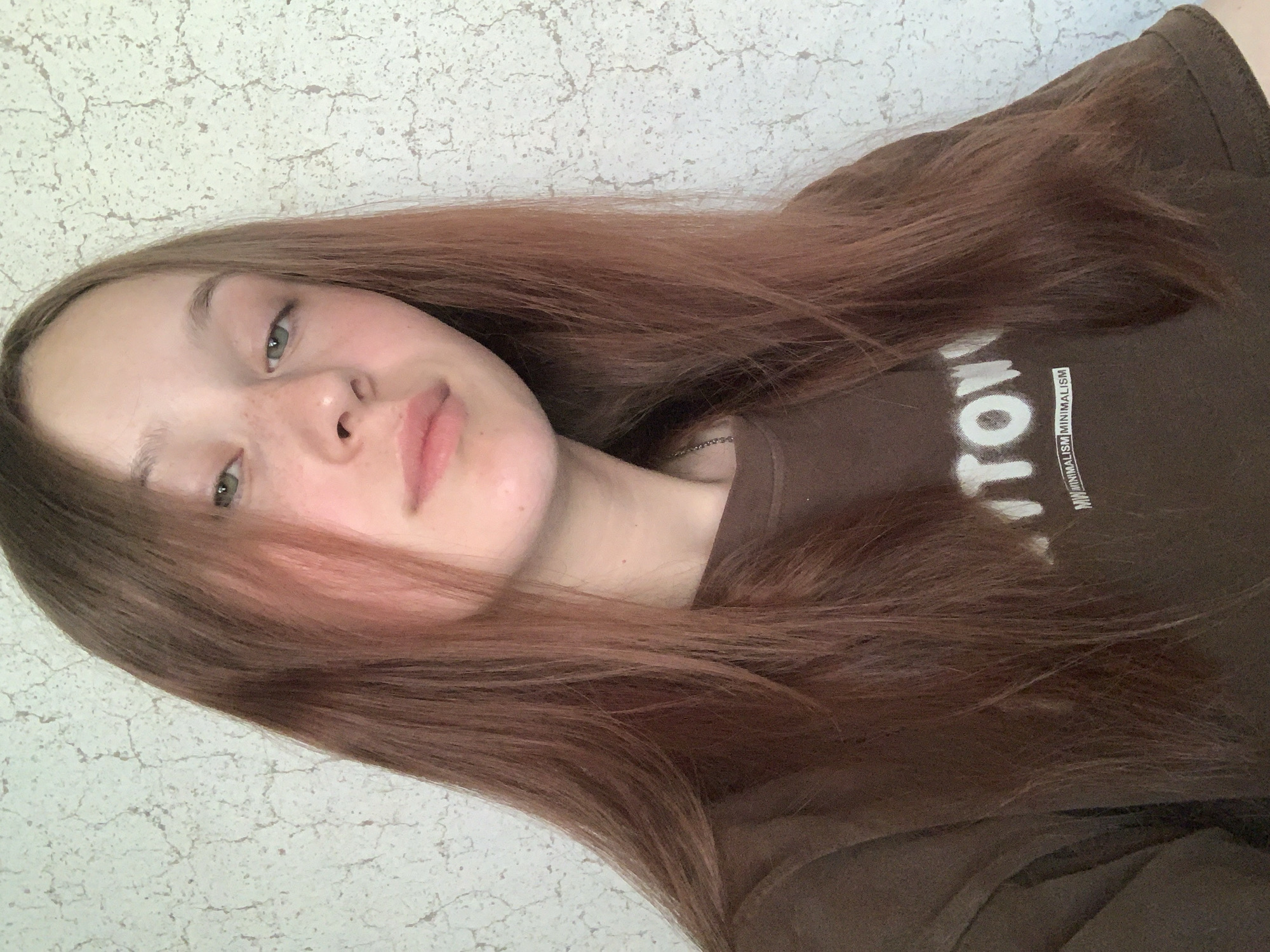 A young woman with long, brown hair is posing indoors against a textured, off-white wall. She is wearing a brown t-shirt with white lettering, part of which reads "MINIMALISM". She has fair skin, freckles, and light-colored eyes. The image is shot from a slightly elevated angle. There is no information available in the image to support any location details, including Фоки, Russia.