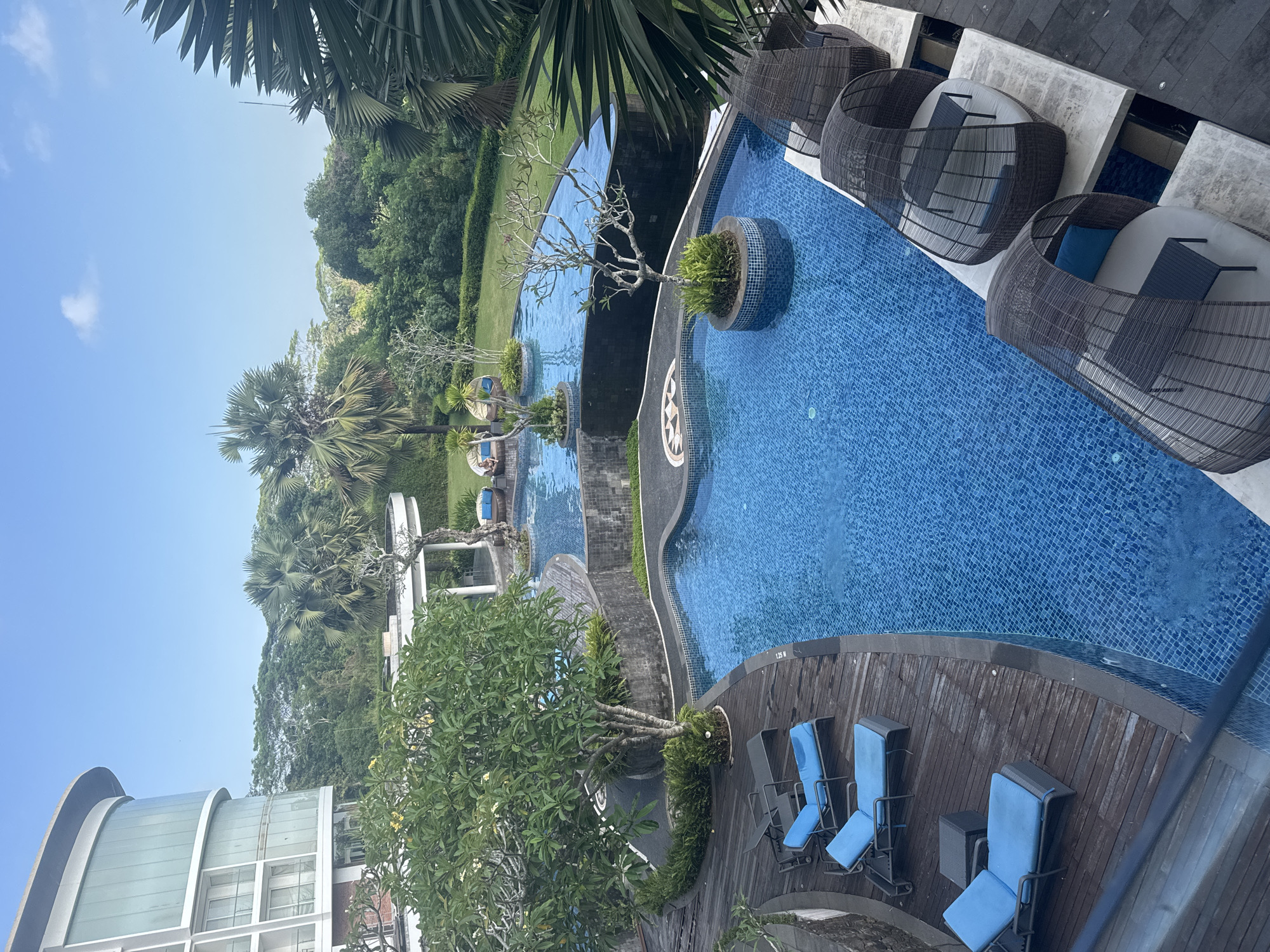 The image depicts a resort swimming pool area in Kuta Selatan, Indonesia. The scene features a multi-level blue tiled swimming pool surrounded by wooden decking and lush greenery. Several blue lounge chairs are arranged on the deck. In the foreground, there are enclosed, cabana-like seating areas with dark wicker exteriors and light-colored cushions. In the background, a modern, curved building with large glass windows is visible, along with palm trees and dense tropical foliage. The sky is clear and blue. There are no people clearly visible in the image.