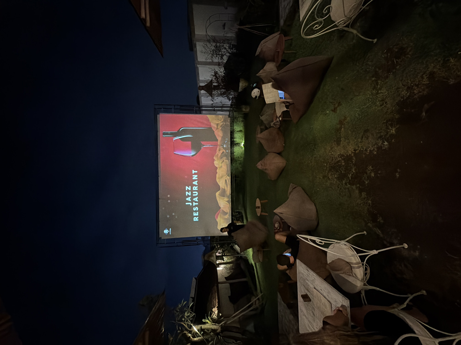 This image depicts an outdoor evening scene at a relaxed dining and entertainment venue, likely located in North Kuta, Indonesia. The central focus is a large projection screen displaying an advertisement for a "JAZZ RESTAURANT." The ad features a stylized image of a wine bottle and a glass set against a warm red and orange background, with the text "JAZZ RESTAURANT" oriented vertically on the right side of the screen.
The setting is a grassy area furnished with numerous casual seating options. Several large, brown, teardrop-shaped beanbag chairs are scattered across the lawn, inviting guests to recline. In addition to the beanbags, some white, ornate metal bistro tables and chairs are visible, particularly on the right side of the frame. A few people are present, seated on the beanbags or at the tables, with one individual appearing to be looking at a phone.
The scene is set at night, evidenced by the dark sky. Illumination comes primarily from the bright projection screen, complemented by subtle ambient lighting that softly highlights the grass, pathways, and surrounding foliage. The atmosphere appears relaxed and casual, suggesting a comfortable outdoor lounge or dining experience.