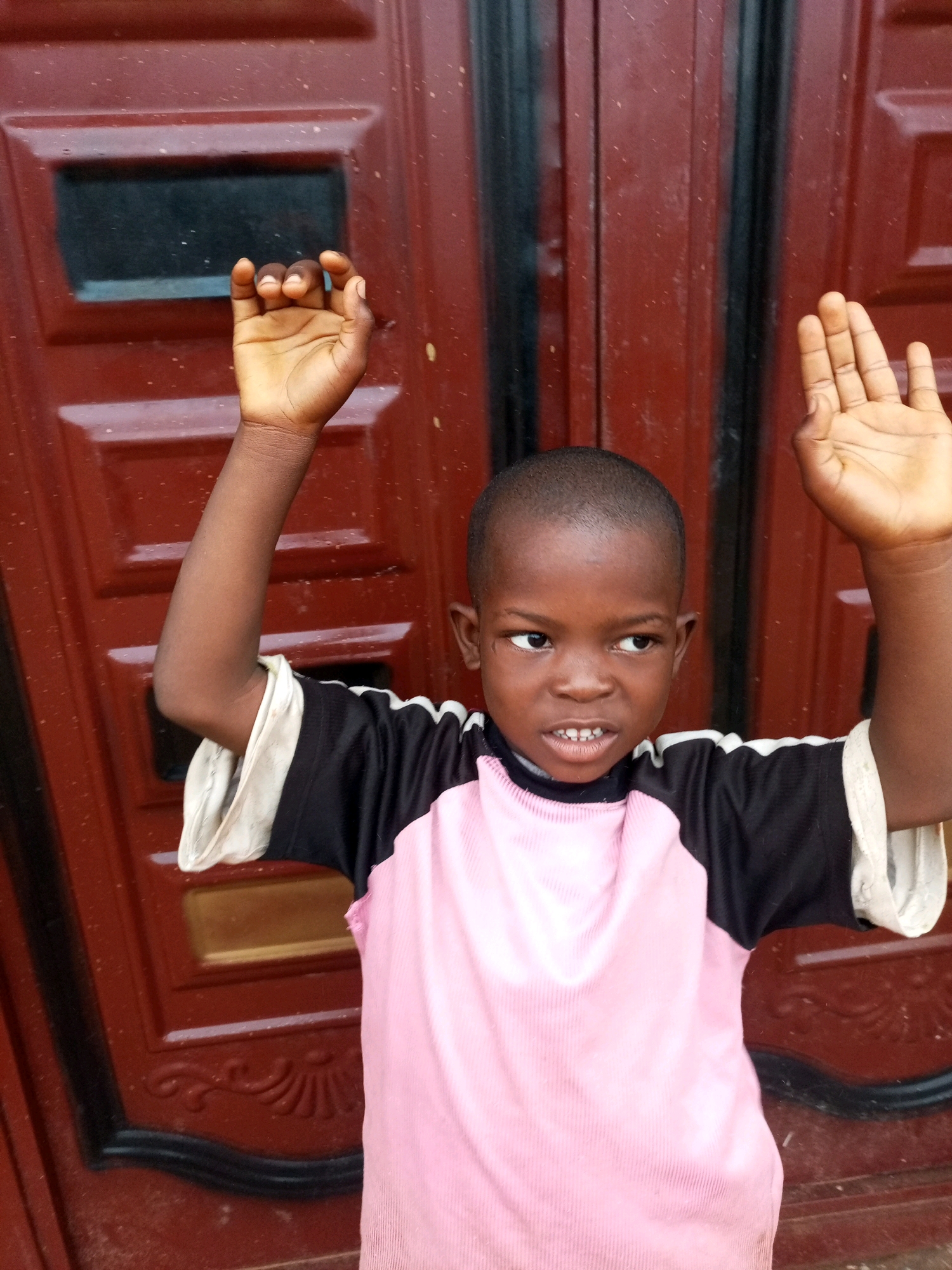A young African boy stands in front of a dark red, paneled door. He wears a pink and black shirt with white trim. His left arm is raised, and his left hand is closed in a fist, fingers curled inward. His right arm is also raised, and his right hand is open, palm facing forward, with fingers spread. He looks to his left with a slight frown and his mouth is slightly open, revealing his teeth. The door has a rectangular window-like panel to the left of the boy's head. The scene appears to be outdoors, possibly in front of a residence.