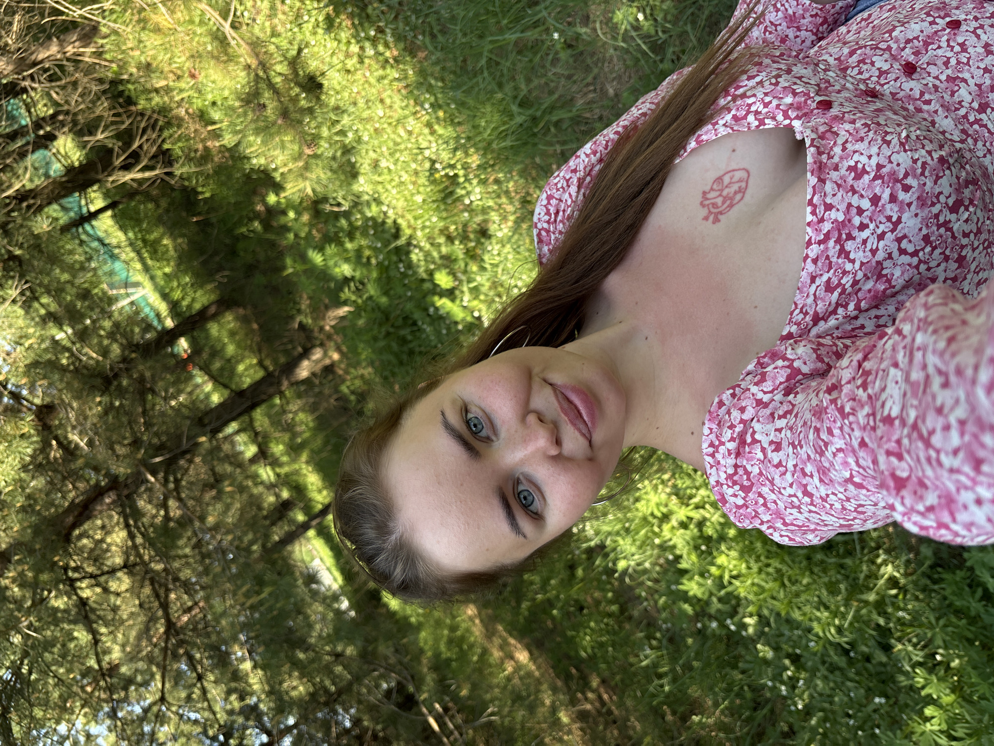 A woman is lying in the grass. She has long brown hair, blue eyes, and is wearing a pink floral dress. She has a red tattoo on her chest. The background consists of trees and grass, suggesting a park or wooded area. Русский Вожой, Russia is not discernible in the image.