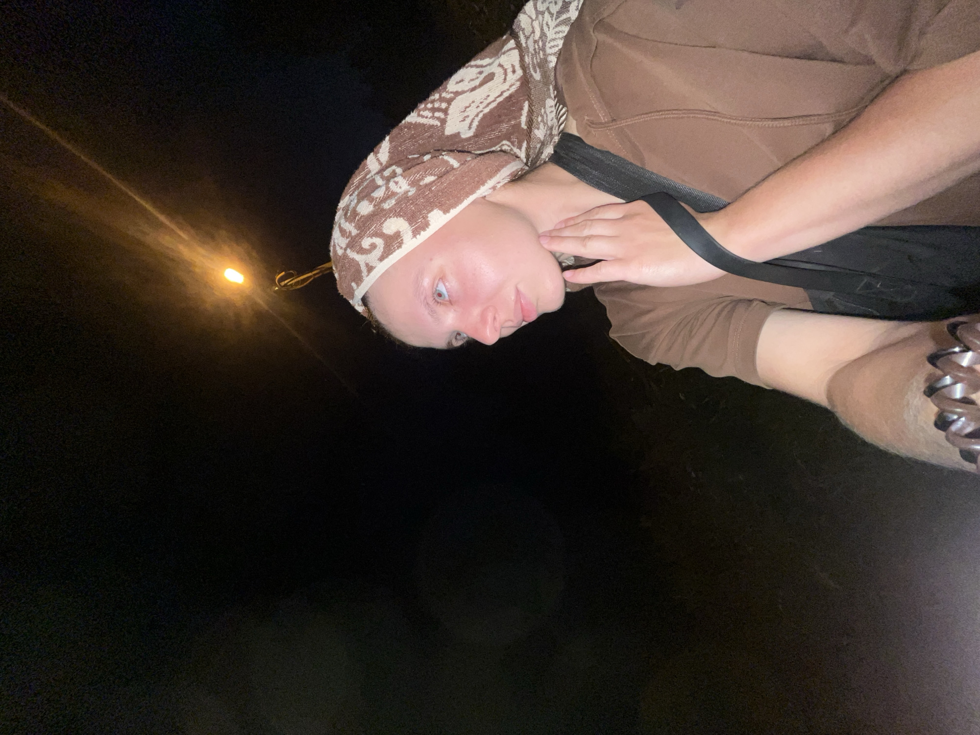 A medium shot captures a young person from the chest up, seen in profile facing left, outdoors at night. They wear a brown hooded sweatshirt and a patterned brown and white headscarf. Their left hand is raised, touching their neck. A black strap, likely from a bag, crosses their chest. The background is dark, with a single light source, possibly a street lamp, visible in the upper right quadrant of the image. The scene is set in the city of Nizhny Tagil, Russia.