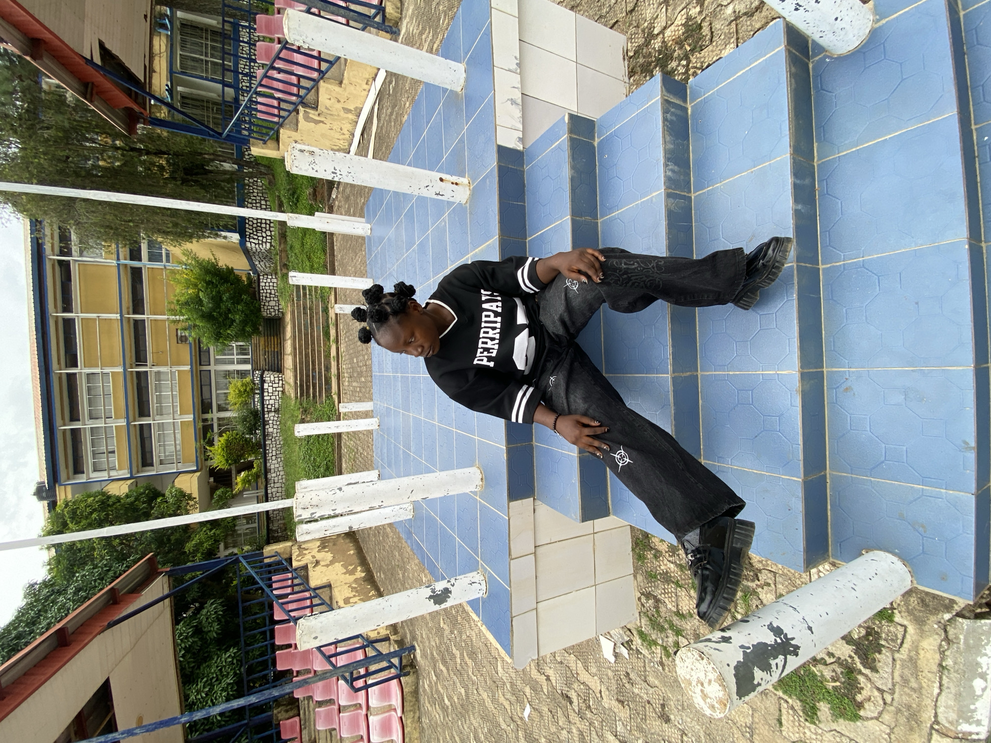 An adult female is sitting on a set of blue-tiled stairs in an outdoor setting. She is wearing a black long-sleeved shirt with white stripes on the sleeves and the word "PERRIPAY" in white lettering across the chest. She is also wearing black baggy pants with a white graphic design and black platform shoes. Her hair is styled in multiple small buns. The stairs are part of a raised platform with white cylindrical pillars surrounding it. In the background, there is a yellow building with multiple windows, a red-roofed structure with pink bleachers, and trees. The overall environment suggests a school or public facility in Zawan, Nigeria.