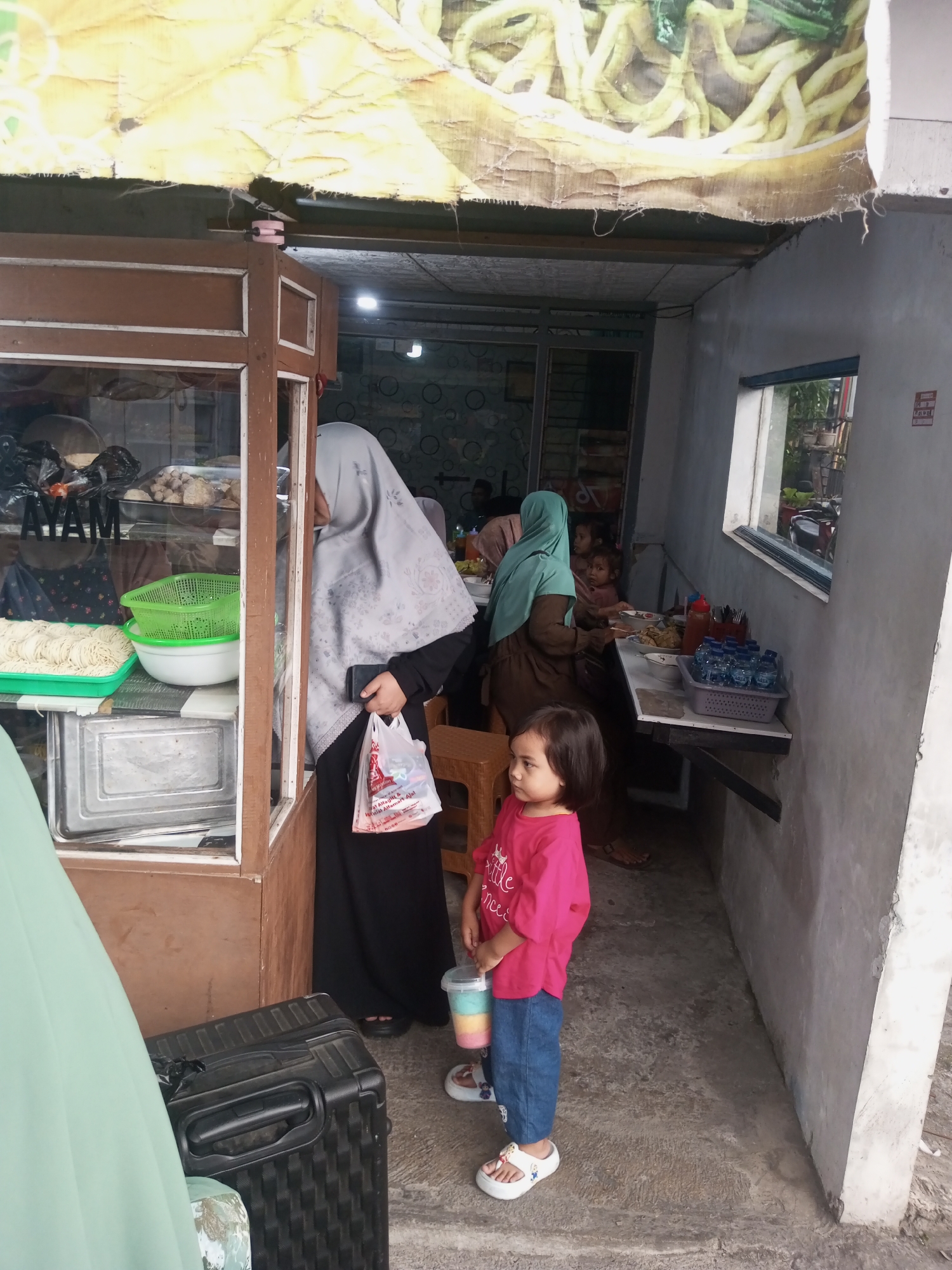 The image depicts a street-side food stall in Pandansari, Indonesia. In the foreground, a young girl wearing a bright pink t-shirt and blue jeans stands holding a colorful drink. To her left, a woman in a grey hijab is partially visible, holding a white plastic bag and a black mobile phone. Behind them, the food stall features a display case with food items, including what appears to be raw noodles and meatballs. Further inside the stall, several people, including women and children, are seated at tables eating. The stall has a rustic appearance with a tarp-covered roof. The ground is concrete, and the walls are plastered. A large black suitcase sits on the ground to the left of the girl.
