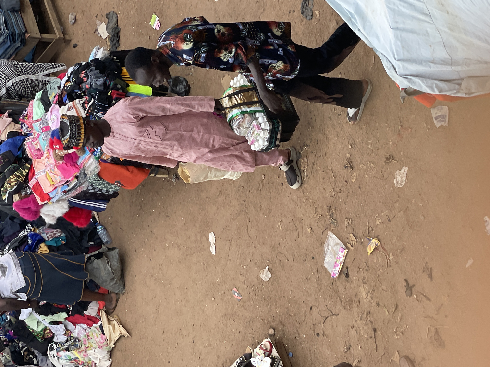 Captured from an overhead perspective, the image portrays a bustling street scene in Unknown, Unknown. The unpaved ground is littered with discarded materials like paper and leaves. Two men are the central figures: one is clad in a pink outfit and a patterned head covering, while the other dons a shirt with floral patterns and black pants. Both are carrying items, including what appears to be a container full of small items. Adjacent to them, a pile of assorted clothes lies on the ground. Additionally, the edge of a white fabric is seen on the right side of the frame.