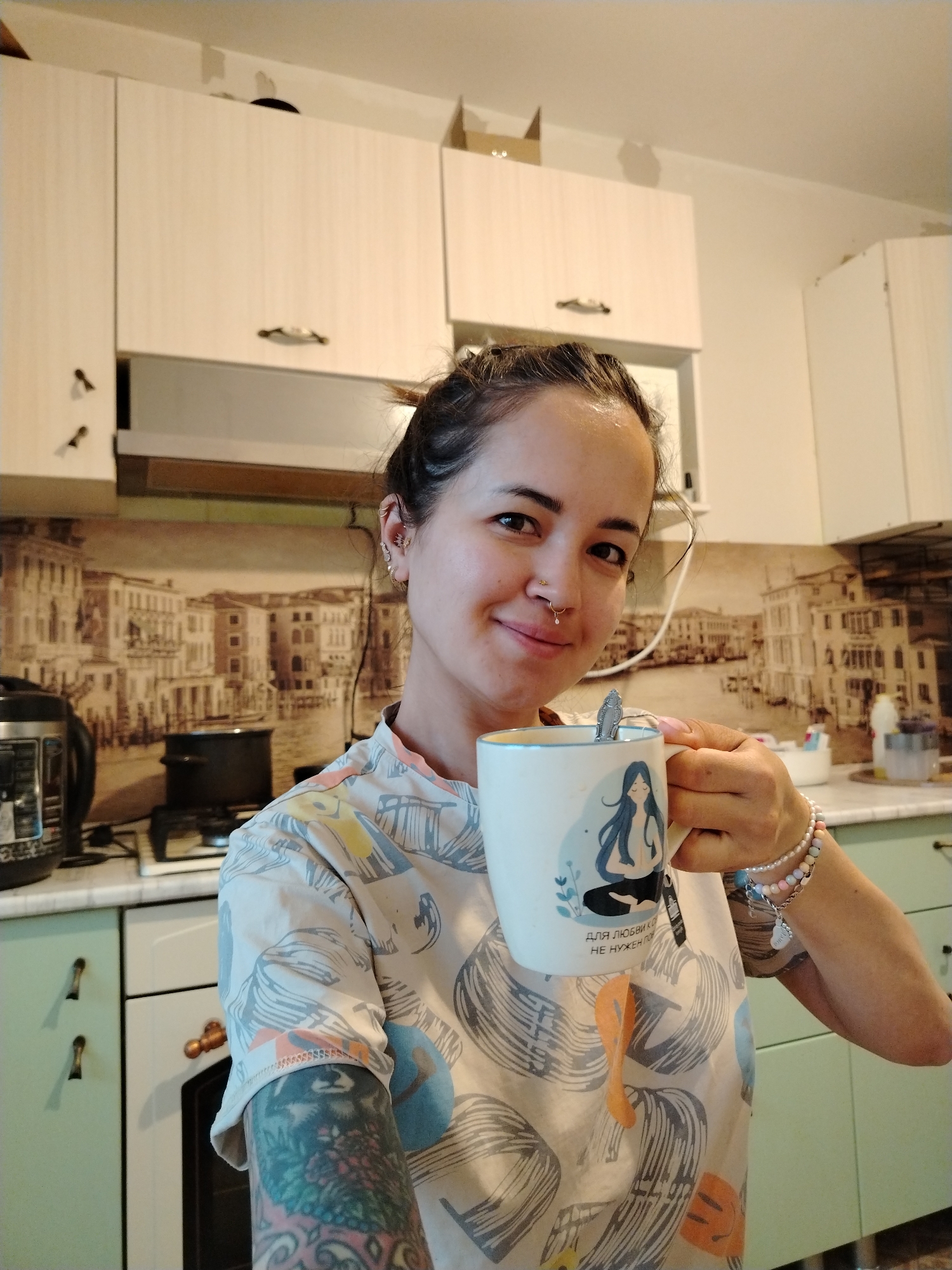 A person is holding a white mug with a blue drawing of a woman in a meditative pose. The person has multiple piercings on their ear and a nose ring. They are wearing a patterned shirt with blue, orange, and grey designs. Their left arm has a tattoo.
The person is in a kitchen. The kitchen has white cabinets, a green countertop, and a backsplash depicting a city scene that resembles Venice, Italy. A multi-cooker and pot are on the counter.