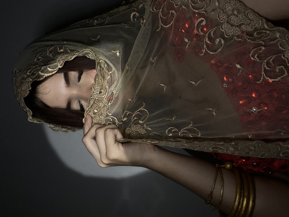 A woman with closed eyes is draped in a translucent, gold-trimmed veil. Her hand, adorned with gold bracelets, partially conceals her face with the fabric. The veil has an embroidered floral pattern and small, decorative stones. The background is a dark gradient, with a light source creating a subtle glow on the left side. The overall effect is one of mystery and elegance.