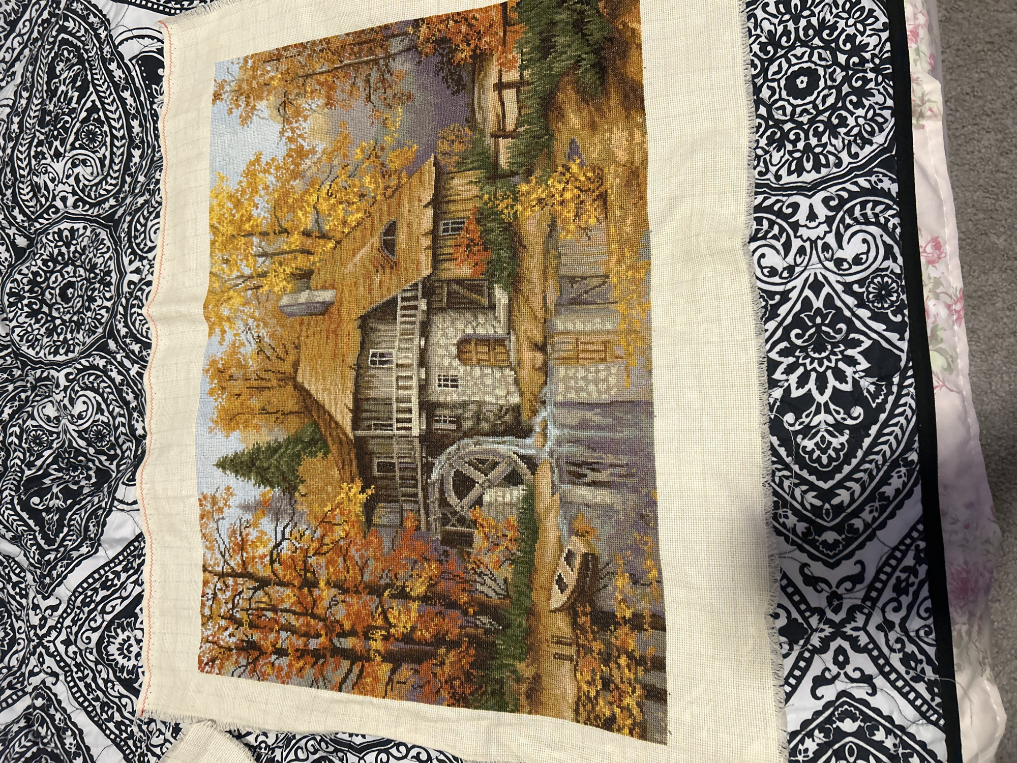 The image shows a partially completed cross-stitch embroidery depicting an autumn scene with a mill house and a water wheel. The embroidery is laid flat on a surface with a black and white patterned fabric underneath. The scene itself features a stone and wood mill house with a thatched roof, surrounded by trees with vibrant orange and yellow foliage. A water wheel is positioned on the side of the mill house, with water flowing from it into a pond. A small boat is visible in the pond, and its reflection is mirrored in the water. The background shows a hazy, autumnal landscape. There are no people visible in the image. The embroidery is unfinished, with the edges of the fabric showing. The overall scene is one of a peaceful, rural setting during the fall season. There is no information in the image to suggest a location in Colorado Springs, United States of America.