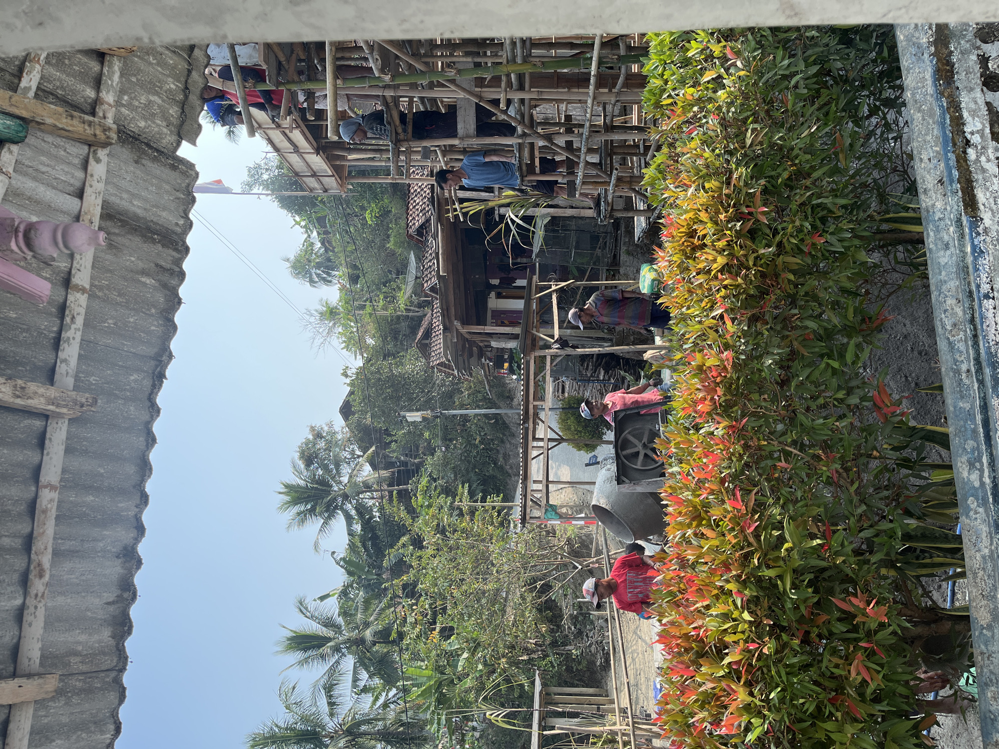 The image depicts a construction site in Kemangguhan, Indonesia. The foreground is dominated by a dense shrubbery with vibrant red and green leaves. Behind the shrubbery, a concrete mixer is visible, alongside several construction workers. One worker, wearing a red t-shirt and a cap, stands near the mixer. Another worker, also in a red shirt, is seen further to the right. In the background, a multi-story bamboo scaffolding is under construction, with several workers on different levels. Tropical vegetation, including palm trees and banana trees, surrounds the site, suggesting a warm climate. Several buildings with tiled roofs are also visible on a gentle slope in the distance. The sky is clear and blue, indicating a sunny day.