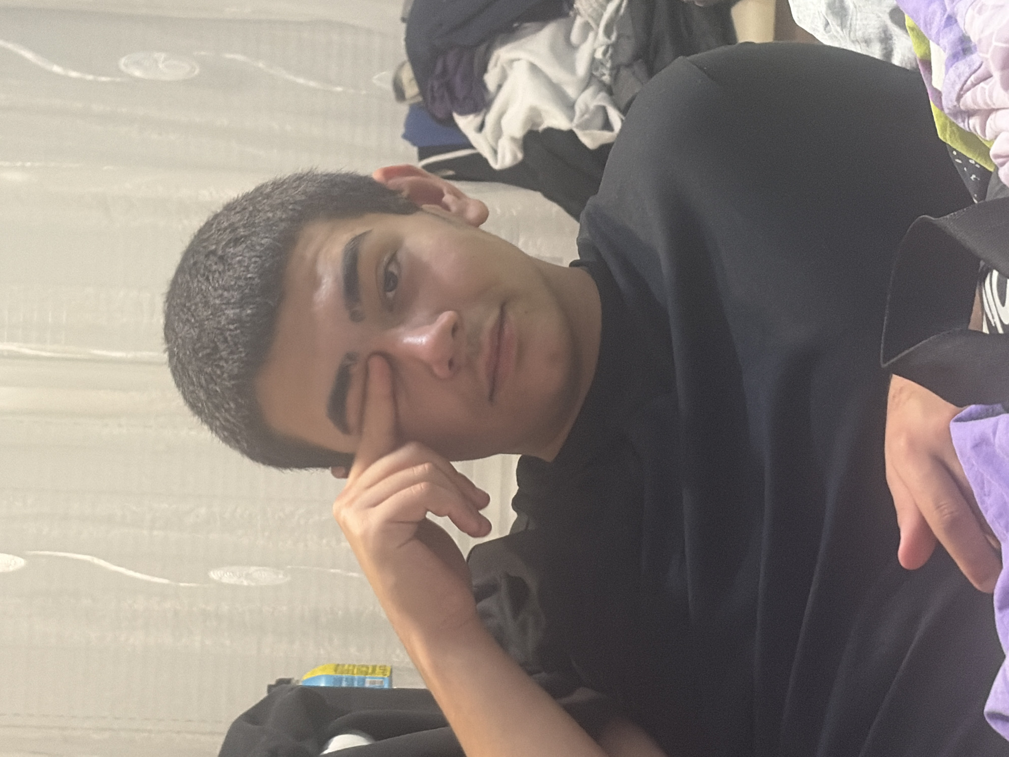 A young male with short, dark hair and dark eyebrows is shown in a medium close-up shot. He is wearing a black t-shirt and is seated, with his left arm raised and his index finger touching his right eye. His gaze is directed towards the viewer. The background consists of a light-colored curtain with vertical, wavy white patterns. To the right of the subject, a pile of folded laundry is visible, including items in white, grey, and purple. The lighting appears to be indoor ambient light. The scene is likely a residential interior, possibly a bedroom or living area. There is no discernible information to place this image specifically in Novi Iskar, Bulgaria.