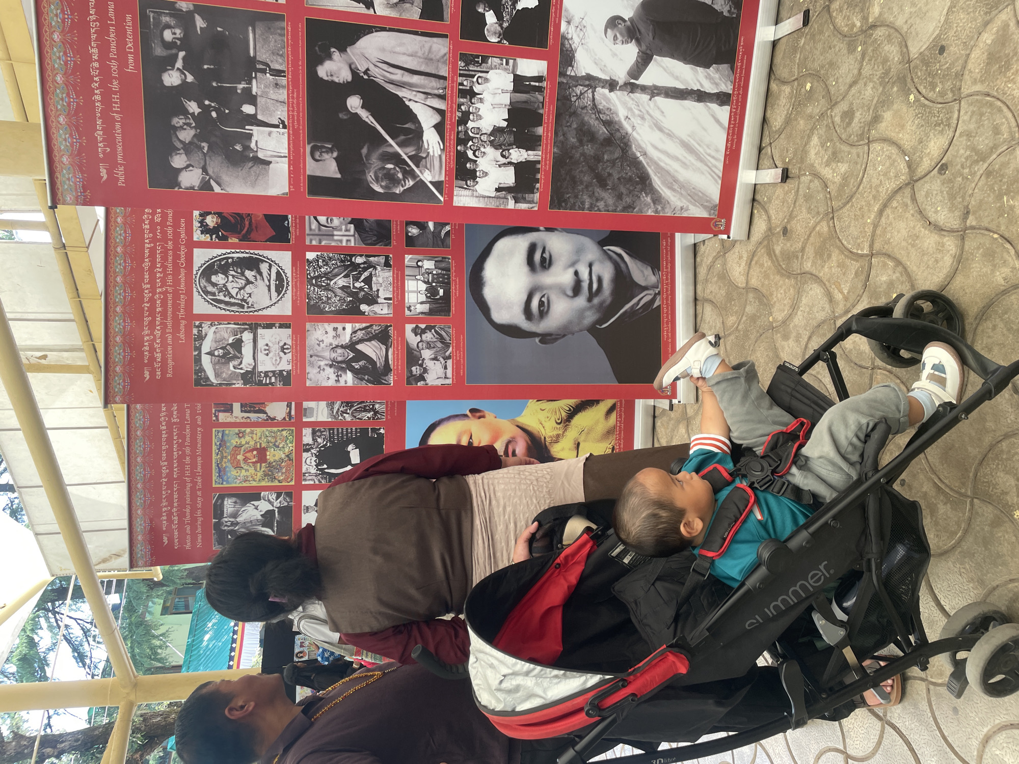 Here's a detailed description of the image:
The image shows a family in front of two vertical red banners with black and white photos and text in Tibetan and English. The banners detail the lives of the 9th and 10th Panchen Lamas, including details of their recognition, travels, and prosecution.
In the foreground, a person is seated on a bench, visible from the shoulders up, wearing a brown and tan robe. To their left, another person is visible from the shoulders up, wearing a dark shirt and gold chain. In front of the people, there is a "summer" brand stroller holding a baby with light skin, wearing a teal shirt, grey pants, and sneakers, with one leg raised in the air. The stroller is black with red trim.
The setting is an outdoor area in the city of Dharamshala, India, recognizable through the presence of traditional Tibetan religious displays and architectural elements. The ground is tiled. A metal-framed awning provides overhead cover.