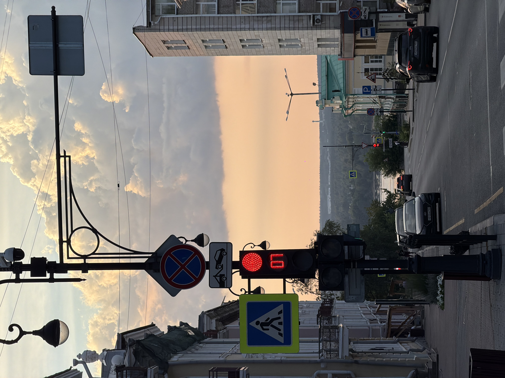 The image captures a street scene in Perm, Russia, with a traffic light displaying red and a countdown timer showing "07". A pedestrian crossing sign is visible on the left, and a no-parking sign is overhead. The street is lined with buildings, and several cars are present, including a black SUV and a black sedan. The sky is filled with dramatic, illuminated clouds, suggesting either sunrise or sunset. In the distance, a river and a forested landscape can be seen, with a tower in the far background.