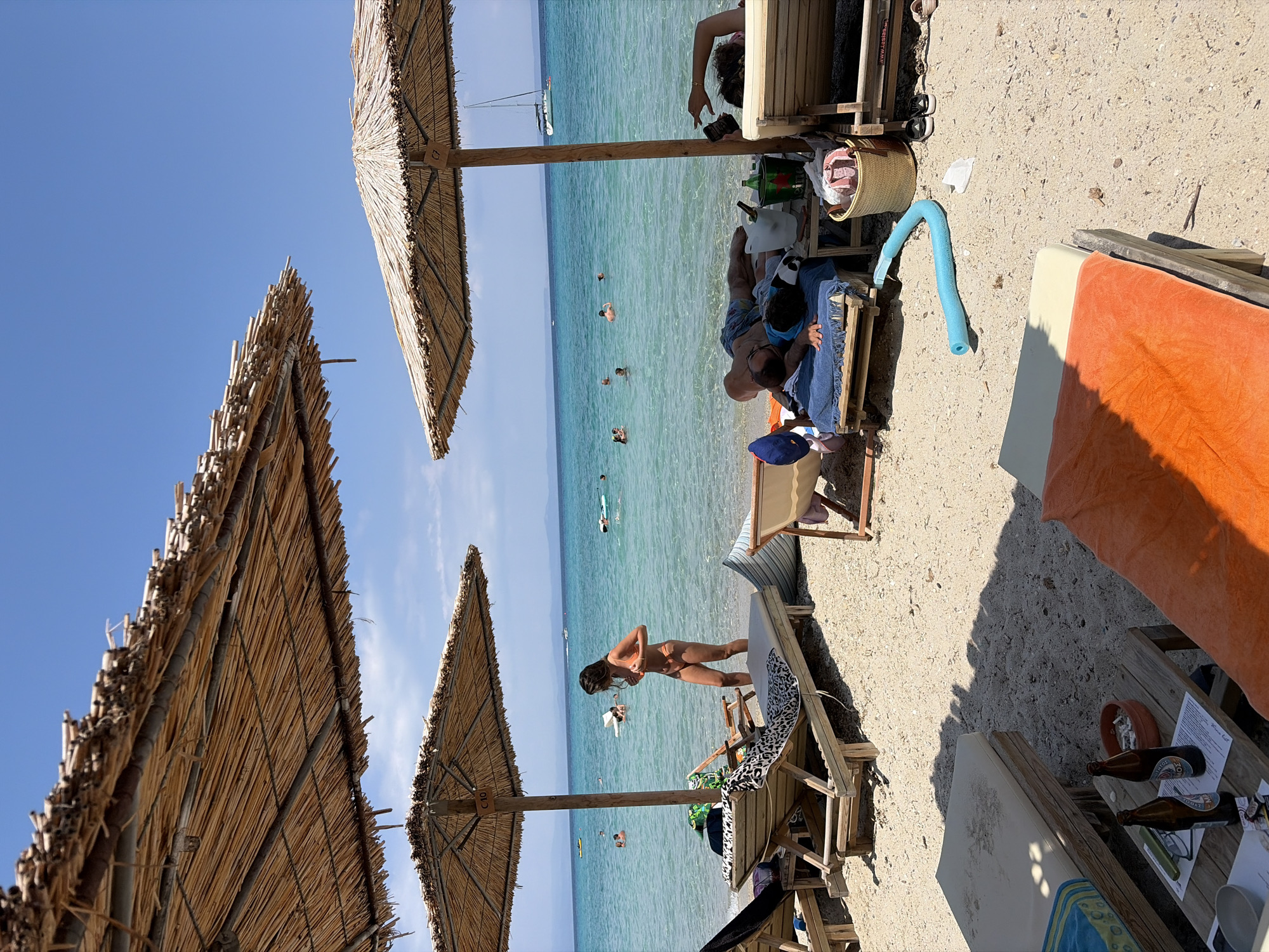 A beach scene under straw umbrellas in Kassandra Municipal Unit, Greece, depicts a serene summer day. The turquoise waters of the Aegean Sea stretch to the horizon, dotted with a few boats and swimmers. In the foreground, several lounge chairs are arranged on the sandy shore.
One woman in a bikini stands near a lounge chair, adjusting her attire, while two men are shown resting on lounge chairs in the shade of an umbrella. To the right, a person's arm is visible, holding a phone. Scattered on the sand are personal items such as towels, a blue pool noodle, a basket, and bottles. The atmosphere suggests a relaxing day at the beach, with people enjoying the sun, sea, and leisure.