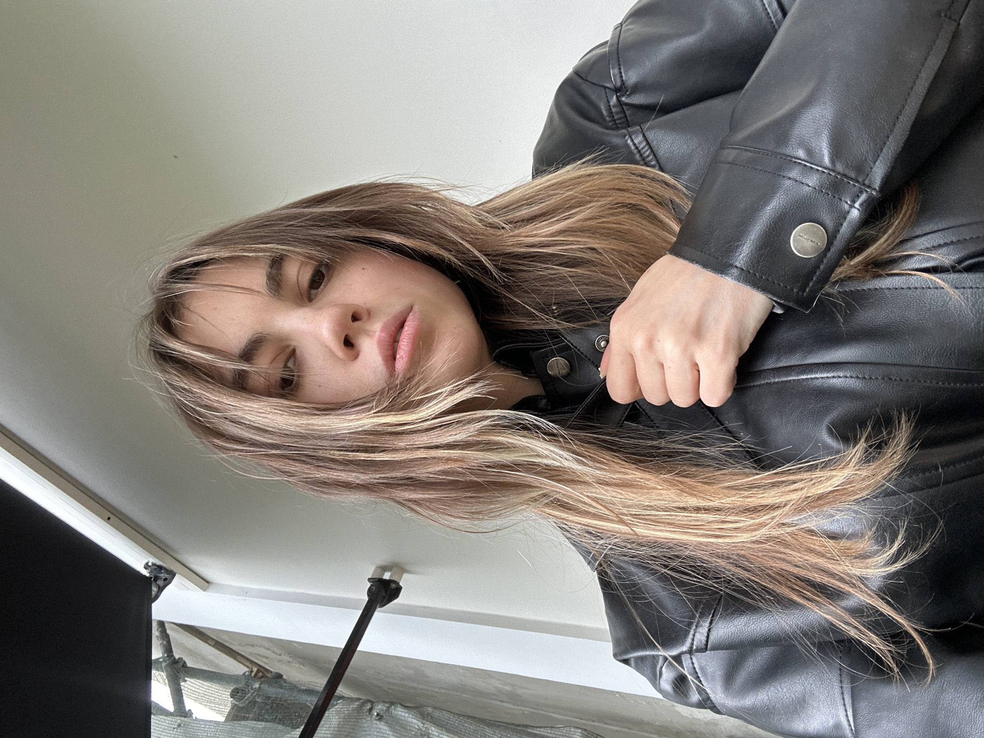 A young woman with long, light brown hair and bangs is shown from a low-angle perspective, looking upwards towards the camera. She is wearing a black faux-leather jacket. Her face is partially obscured by her hair, and she has a neutral expression. The scene appears to be indoors, possibly near a window, as evidenced by the bright, diffused lighting and a glimpse of what looks like scaffolding and netting in the background, suggesting construction or renovation work. The environment is minimalist, with white walls or ceilings dominating the upper part of the frame. A thin, metal pole with a black fixture is visible in the lower part of the image. A small, dark speck, possibly a fly or dust, is visible on the white surface in the upper left quadrant. The image appears to be a selfie, taken from a downward angle. There is no visible text in the image.