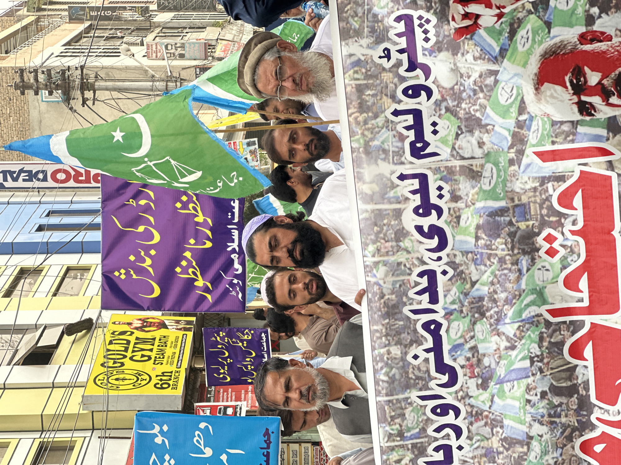 The image depicts a political rally in Quetta City Tehsil, Pakistan. The foreground features a banner with Urdu text, partially obscured by several men. One man in the center wears a grey vest over a light-colored shirt and has a grey beard and mustache. To his right, another man has a beard and wears a white cap and a white tunic. Behind them, more men are visible, some holding flags.
A prominent flag, green with a white crescent moon and star, and a scale of justice emblem, is visible towards the right. Other flags, also green and white, are scattered throughout the crowd. In the background, buildings line a street, adorned with various signs, including a "Gold's Gym" sign and other advertisements in Urdu. The environment appears to be an urban setting with overhead wires and power poles. The overall atmosphere suggests a public gathering or demonstration.