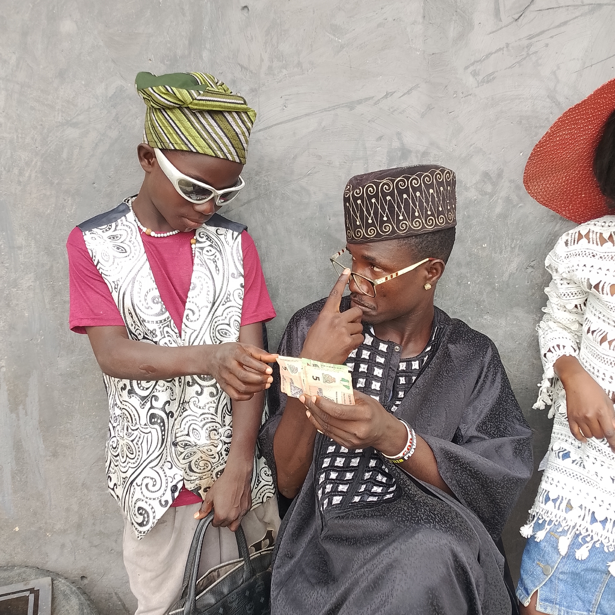 In Ado, Nigeria, two boys and a young woman stand against a textured gray wall. The boy on the left, wearing a green and white striped headwrap, white sunglasses, a maroon t-shirt, and a white patterned vest, looks down at his hands. He holds a black handbag with a silver zipper and a silver clasp. The boy in the center, dressed in a dark cap with intricate gold embroidery, gold-rimmed glasses, and a black garment with a white checkered pattern, looks towards the boy on the left while holding several folded banknotes in his hands. The young woman on the right, partially visible, wears a wide-brimmed red hat and a white lacy top over denim shorts. The scene appears to be an outdoor urban setting.