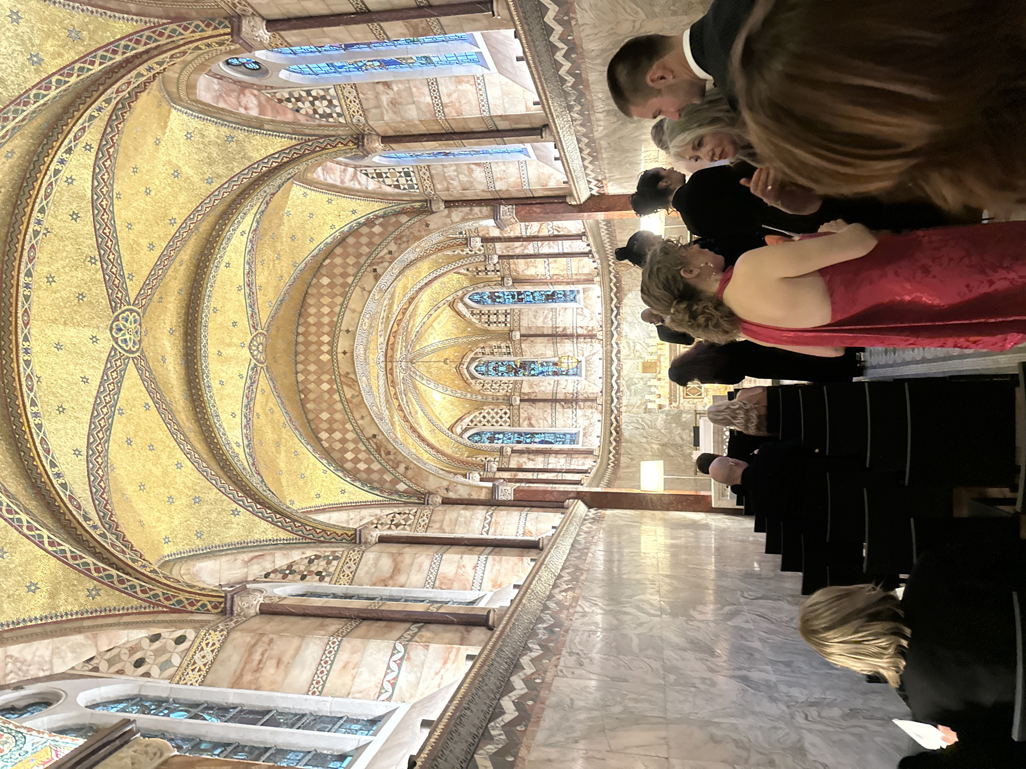 Captured inside a cathedral, the image presents an interior scene filled with people attending an event, likely a wedding or religious service. The architecture is characterized by intricate mosaics adorning the arched ceiling, and stained glass windows are nestled in the walls.
The people are dressed in formal attire, which suggests the solemnity of the occasion. Some are seated in rows of chairs, while others gather in smaller groups, possibly conversing.
The environment exudes an aura of reverence and history.
The location is likely Westminster Cathedral in London, United Kingdom, based on the architectural style and intricate mosaics.