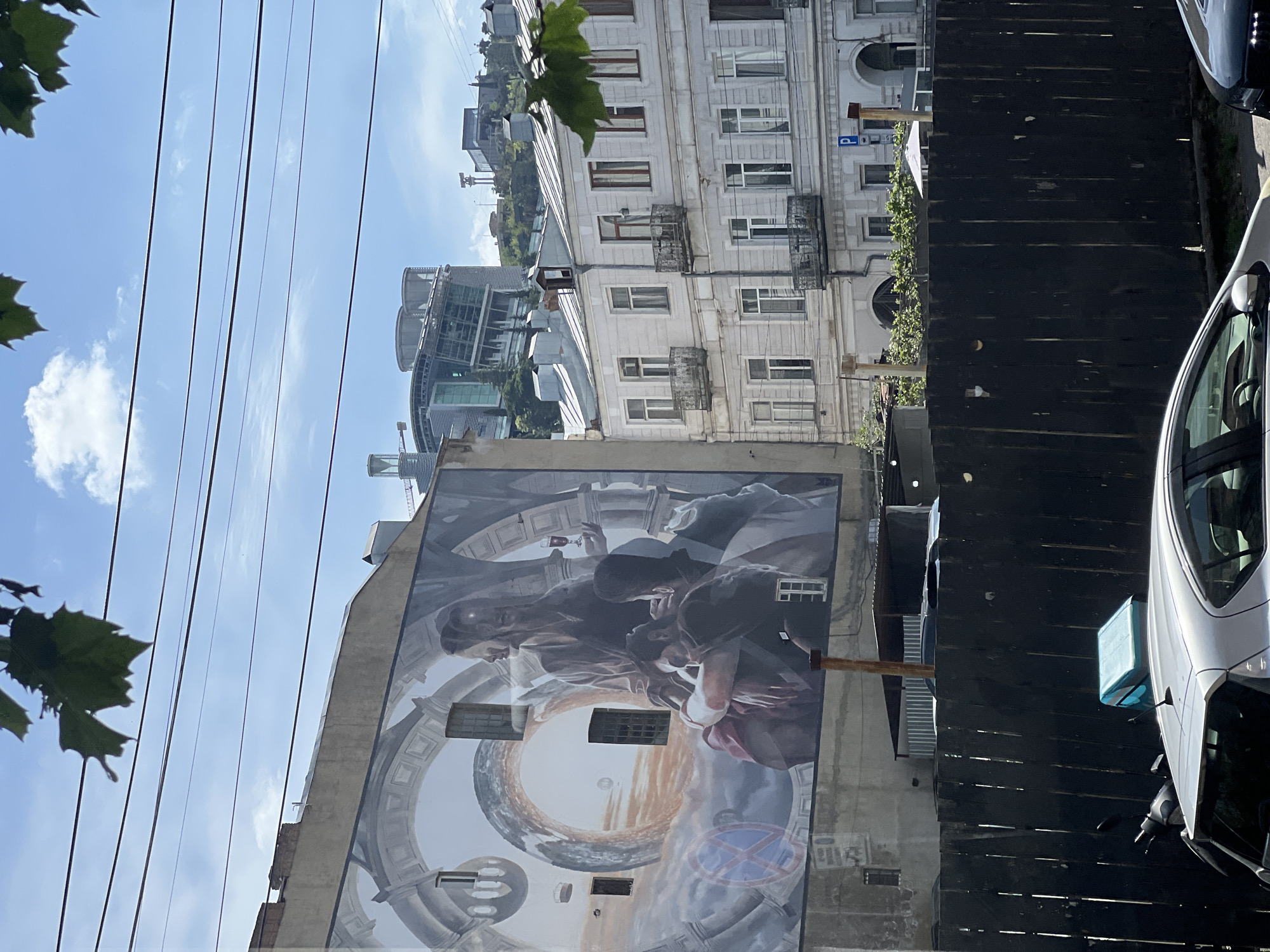 The image is a street-level shot in Tbilisi, Georgia, depicting a building with a large mural on its facade. The mural features figures reminiscent of classical paintings, set against a background of architectural motifs and abstract shapes. Adjacent to the mural building is an older structure with white facade, multiple windows, and small balconies. A dark, wooden fence runs along the street, partially obscuring the lower portion of the buildings. A white car is parked at the right edge of the frame, next to the fence. Overhead, power lines stretch across the sky. Leaves are visible in the left corner, and other buildings, including a modern glass structure, can be seen further back, suggesting an urban setting. The sky is blue with light clouds.