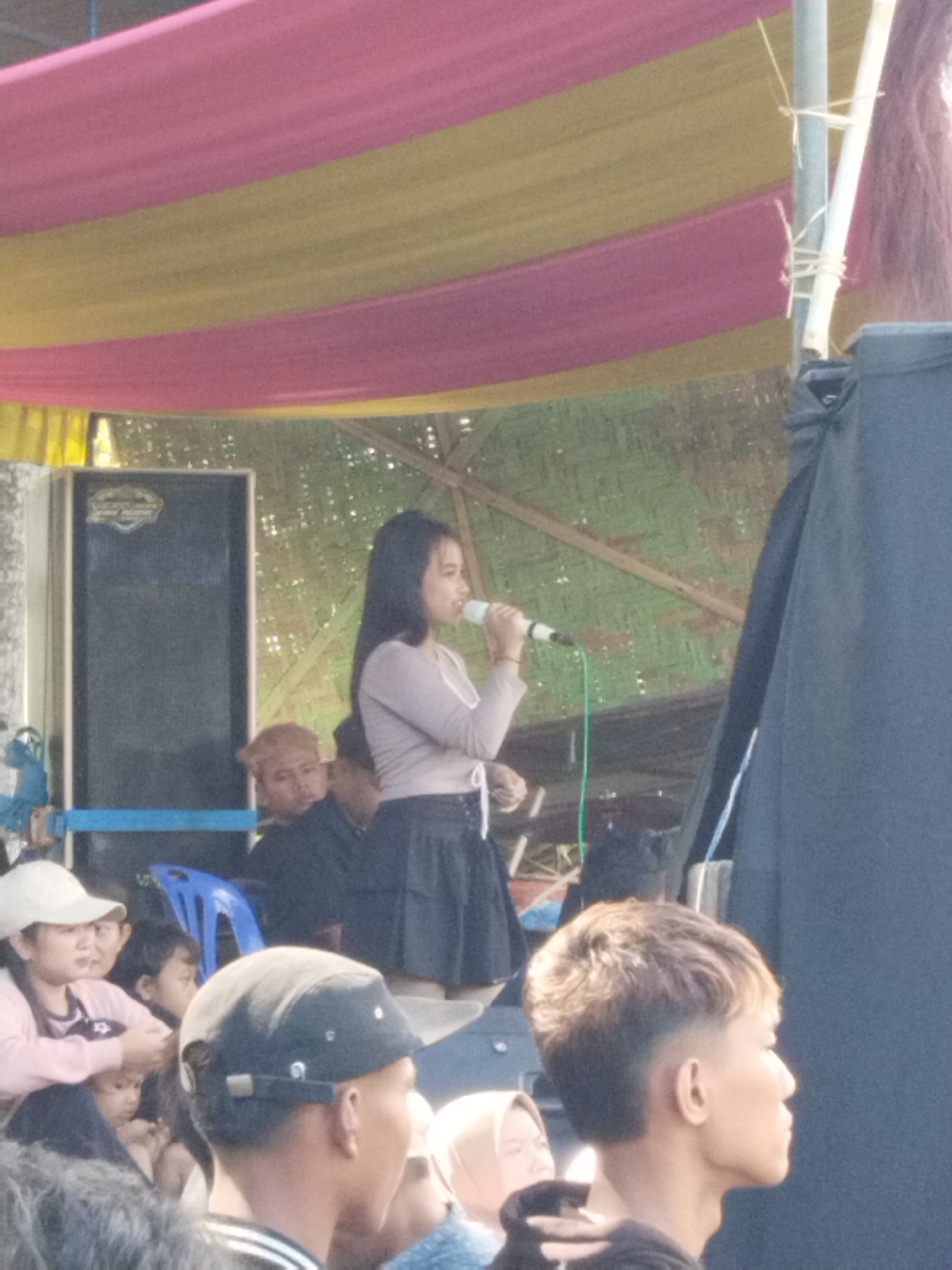A woman is singing on stage in Pendem, Indonesia. She is wearing a long-sleeved shirt and skirt and holding a microphone. A crowd of people is watching her. There is a speaker on stage and a backdrop of woven material. Pink and yellow fabric is hanging over the stage.