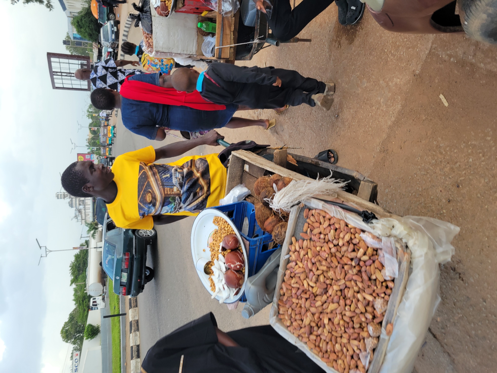 A group of people are gathered around a street vendor's cart in what appears to be Jos, Nigeria. The scene is set on a dirt and asphalt road, with a variety of vehicles, including cars and what looks like a tuk-tuk or auto-rickshaw, visible in the background. The sky is overcast, suggesting a cloudy day.
The main subjects are several individuals, including a vendor actively selling goods from his cart. He is wearing a bright yellow t-shirt with a graphic print. Another person, wearing a black outfit and a red sash over their shoulder, is interacting with someone at the cart. A young person in a dark outfit with a blue collar is also present. The vendor's cart is laden with various food items, including what appear to be dates or similar dried fruits in a large bin, coconuts, and a bowl containing smaller nuts and some white, sliced items.
The activity appears to be a typical street-side transaction, with people browsing and purchasing from the vendor. The overall atmosphere seems bustling and casual, consistent with a marketplace or roadside vending area. There is no visible text in the image that can be clearly read and interpreted.