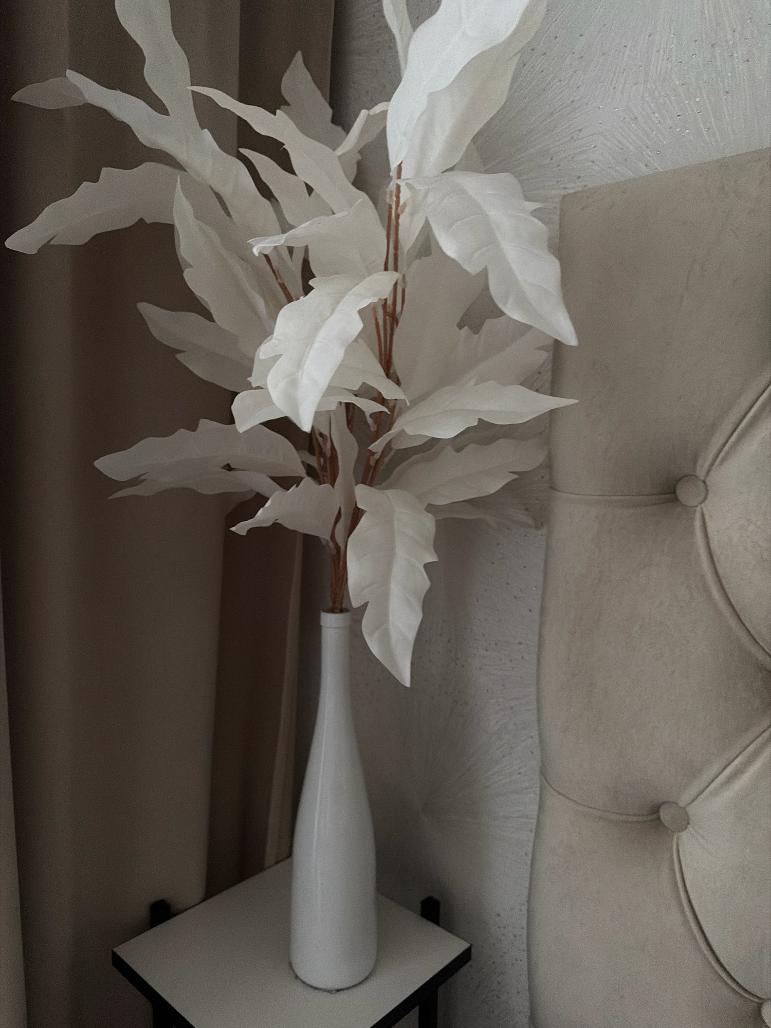 A tall, slender white vase holds a branch with large, white, artificial leaves. The vase sits on a small, white side table with black trim, which is positioned to the left of a cream-colored, tufted headboard. Behind the vase and table, a floor-length, neutral-toned curtain is visible. The headboard has button tufting and is textured with a subtle, raised pattern. The overall scene is minimalist and appears to be indoors, possibly in a bedroom. The location in the city of Otradnaya, Russia is not discernible from the image itself.