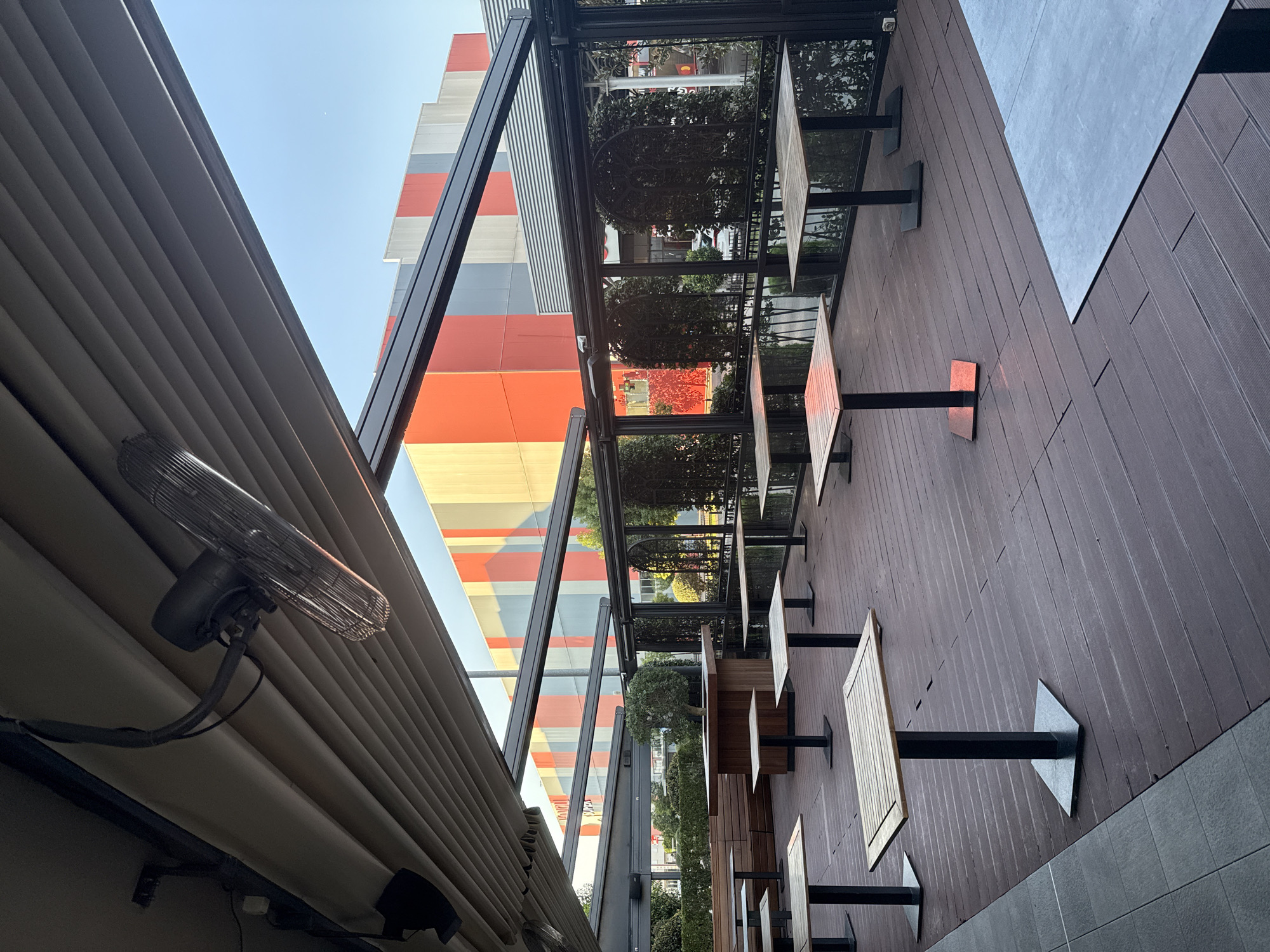 This is a medium shot, outdoor photograph of a covered patio area with tables and chairs, situated in the city of Unknown. The patio is furnished with several small, square wooden tables with black metal bases, and some of these tables are accompanied by black metal chairs. The flooring of the patio is made of dark wooden planks.
The patio is covered by a retractable roof structure with horizontal louvers. Hanging from the roof structure are two black industrial fans. In the background, a large, modern building with a facade featuring vertical stripes of orange, grey, and white is visible. Some greenery, including bushes and a small tree, is present on the patio and beyond the glass enclosure. The sky above is clear and blue. The image appears to have been taken during daylight, possibly late afternoon, as indicated by the warm lighting on some of the tables. No people are visible in the scene.